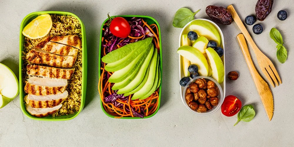 A set of three bento boxes with grilled chicken on quinoa, shredded carrots and purple cabbage with cherry tomato and sliced avocado, apple slices with blueberries, and a small bowl of hazelnuts, on a light gray surface with scattered blueberries, cherry tomatoes, basil leaves, and wooden fork and spoon.