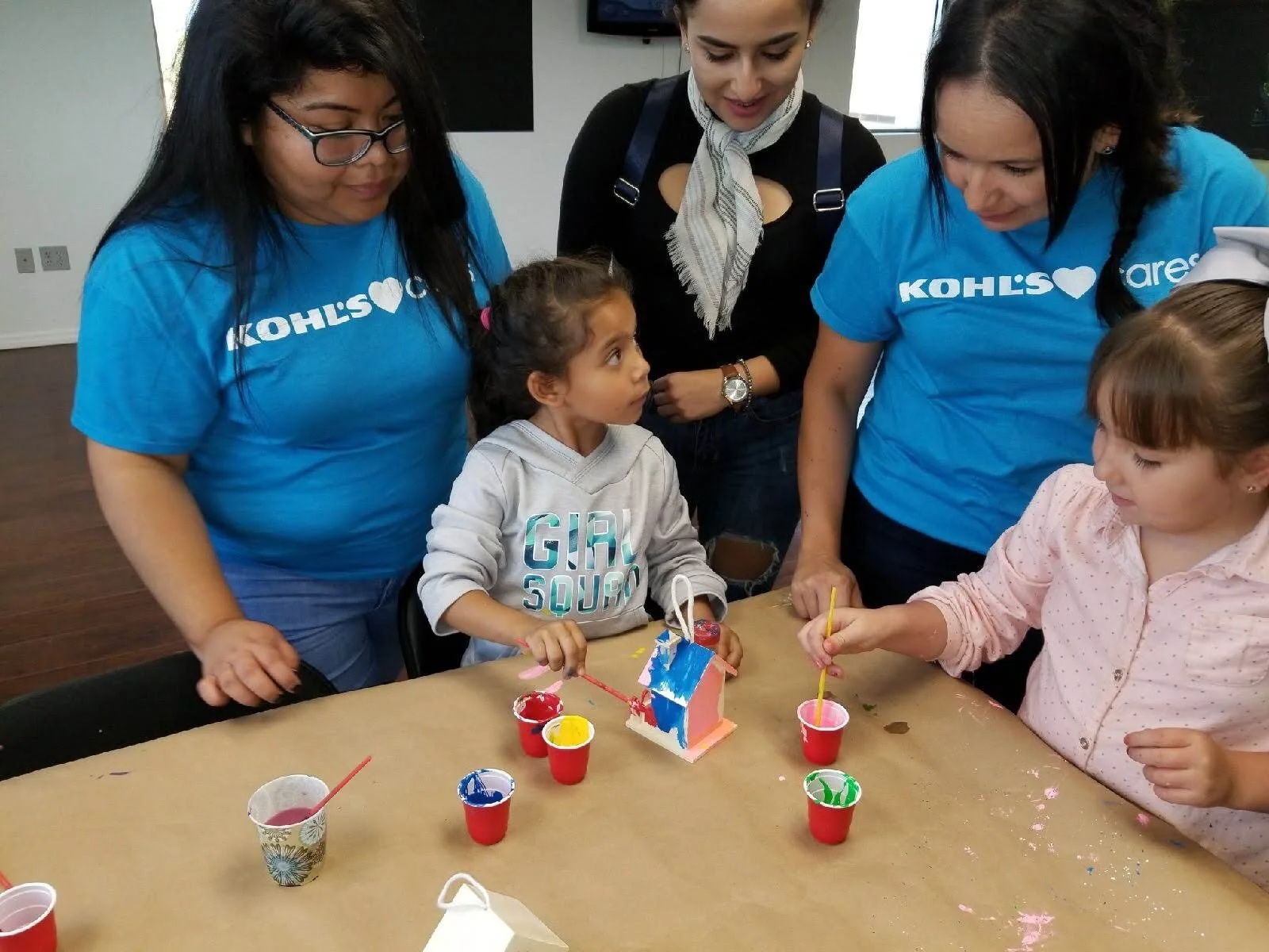 Two young girls are sitting at a table painting a small wooden house with bright colors, while helpers watch and interact. The women are wearing matching blue T-shirts are word 'KOHLS' representatives.