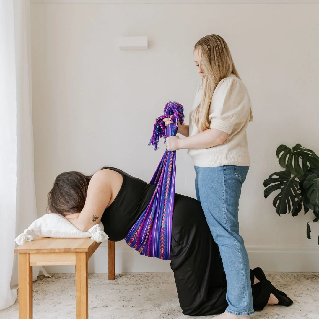 A woman is lying face down on a small wooden table with her head resting on a pillow, while another woman stands behind her, pulling a colorful, woven hammock-like cloth around her.