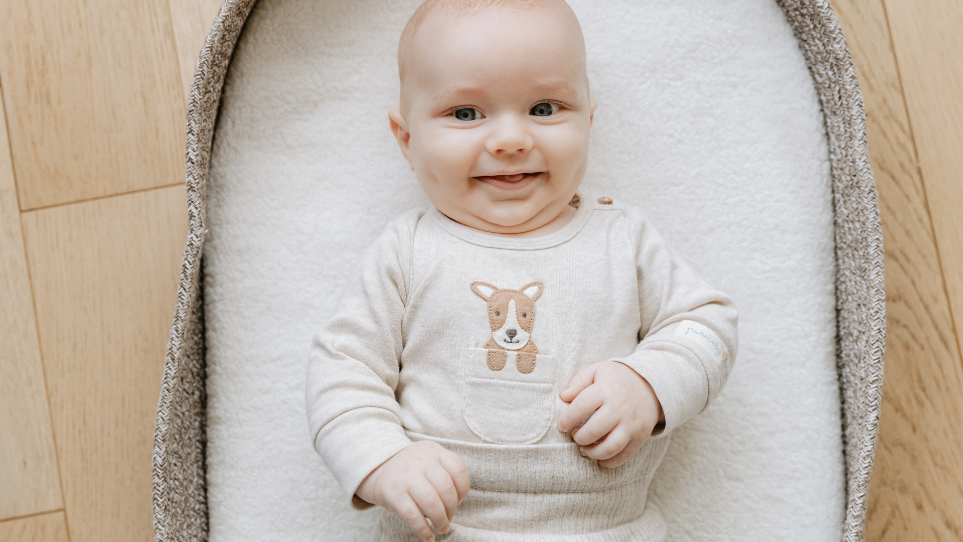 A smiling baby with blue eyes lying on a soft, white blanket inside a cradle, wearing a beige outfit with a dog face embroidery on the chest.