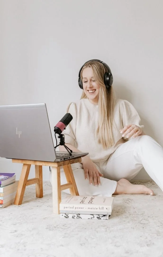 A woman with long blonde hair, wearing headphones and a light sweater, sitting barefoot on the floor next to a small wooden table with a microphone and an open laptop. She is smiling and appears to be recording a podcast or video. There are books stacked on the floor around her, one titled 'period power' by Maisie Hill.