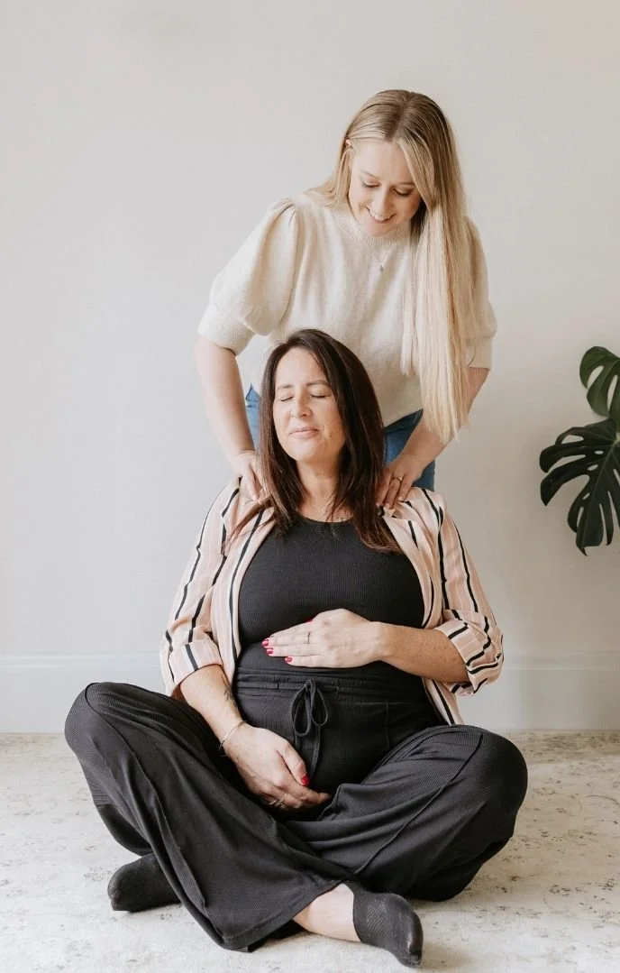 A woman pregnant woman sitting cross-legged on the floor with her hand on her belly, while another woman stands behind her, gently supporting her shoulders in a soothing manner.