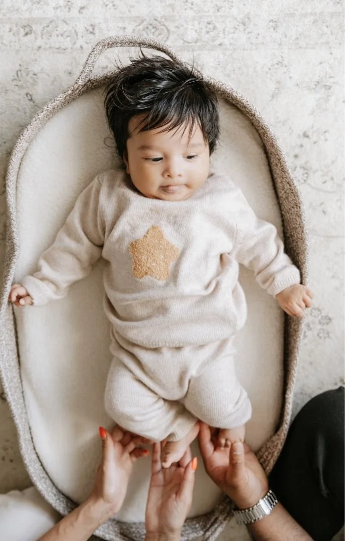 A baby lying on a soft, oval-shaped cushioned changing pad, wearing a light beige outfit with a star pattern, being gently held by an adult's hands.