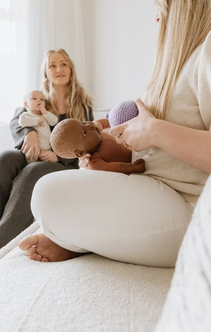 A woman is breastfeeding a baby while sitting on a bed, with a woman and a baby sitting in the background in a well-lit room.