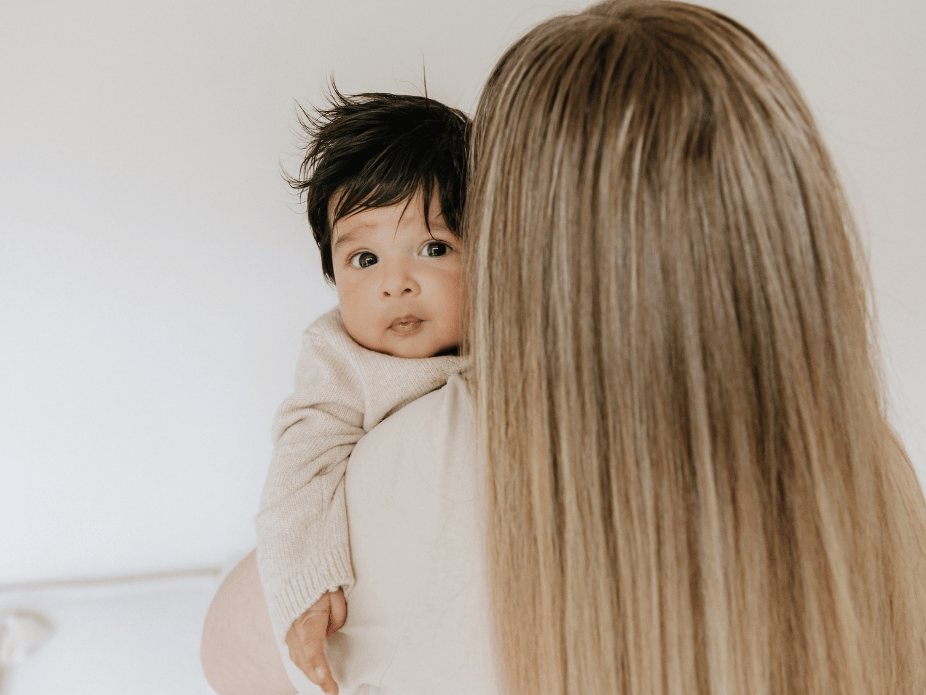 A woman with long blond hair holding a baby with dark hair and big eyes, looking over her shoulder.