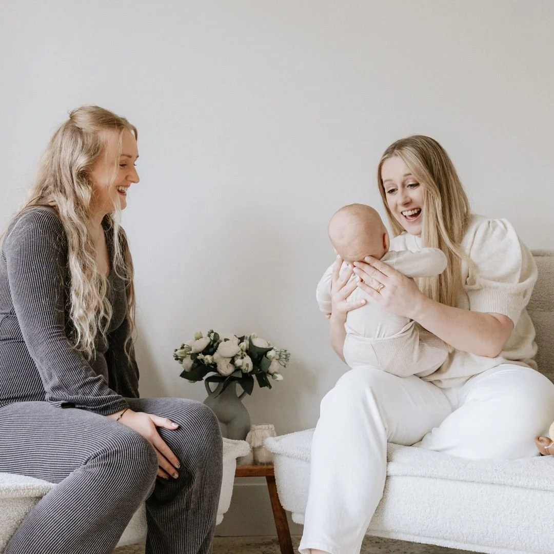 Two women, one with long blonde hair laughing and sitting on the left, and the other with long blonde hair holding a baby on the right. They are in a cozy room with minimal decor, including a wooden table with a vase of flowers.