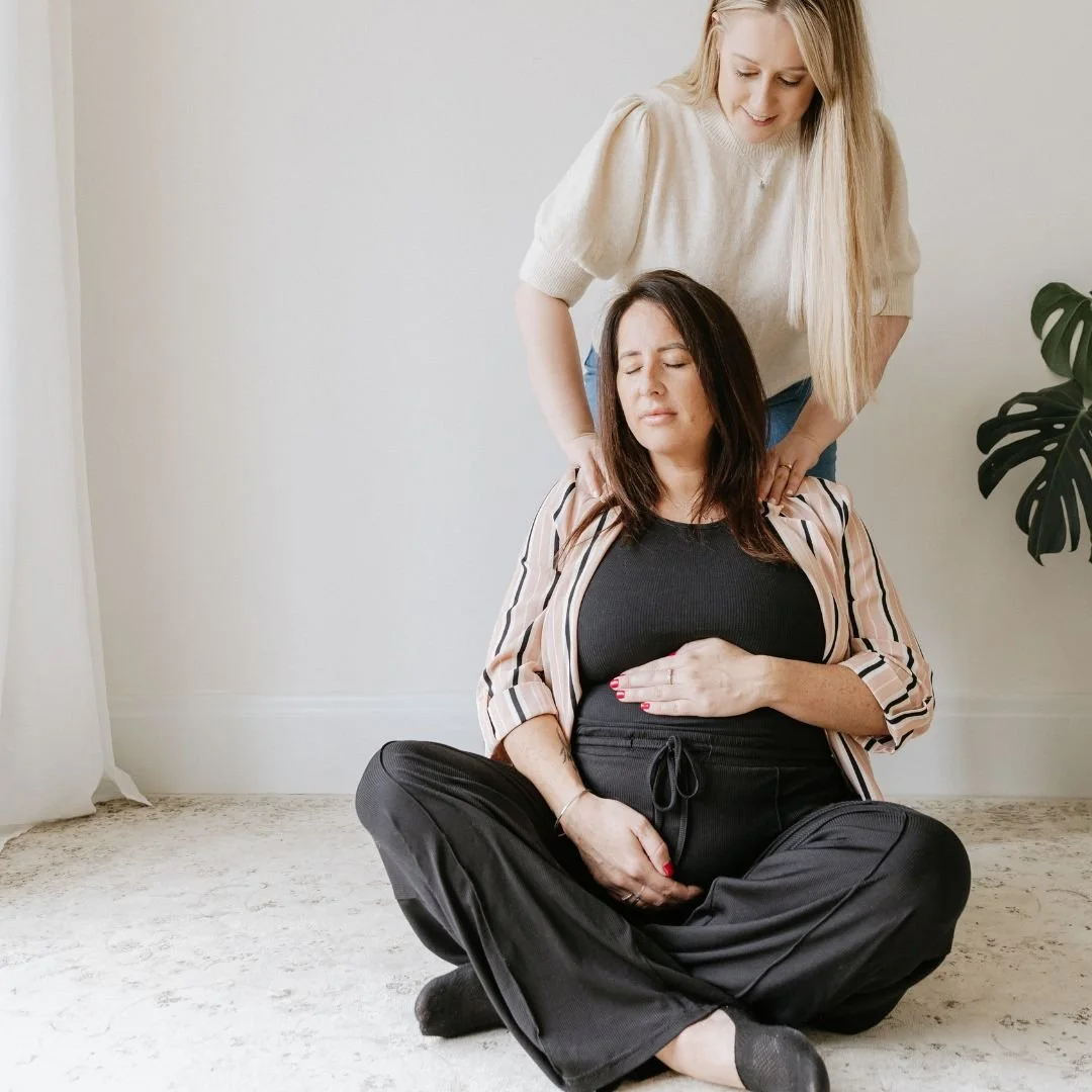A woman sits cross-legged on the floor with her eyes closed, placing one hand on her stomach. She appears to be practicing meditation or relaxation, while another woman stands behind her, gently massaging her shoulders. The setting is a simple room with light-colored walls and a green plant in the background.