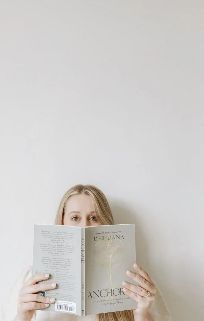 Woman with long hair holding a book titled 'Anchor' by Deb Dana, partially covering her face, against a plain white wall.