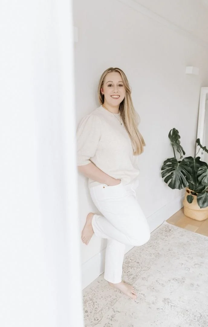 A woman with long blonde hair wearing a cream-colored top and white pants, standing barefoot indoors against a light-colored wall with a large green potted plant nearby.