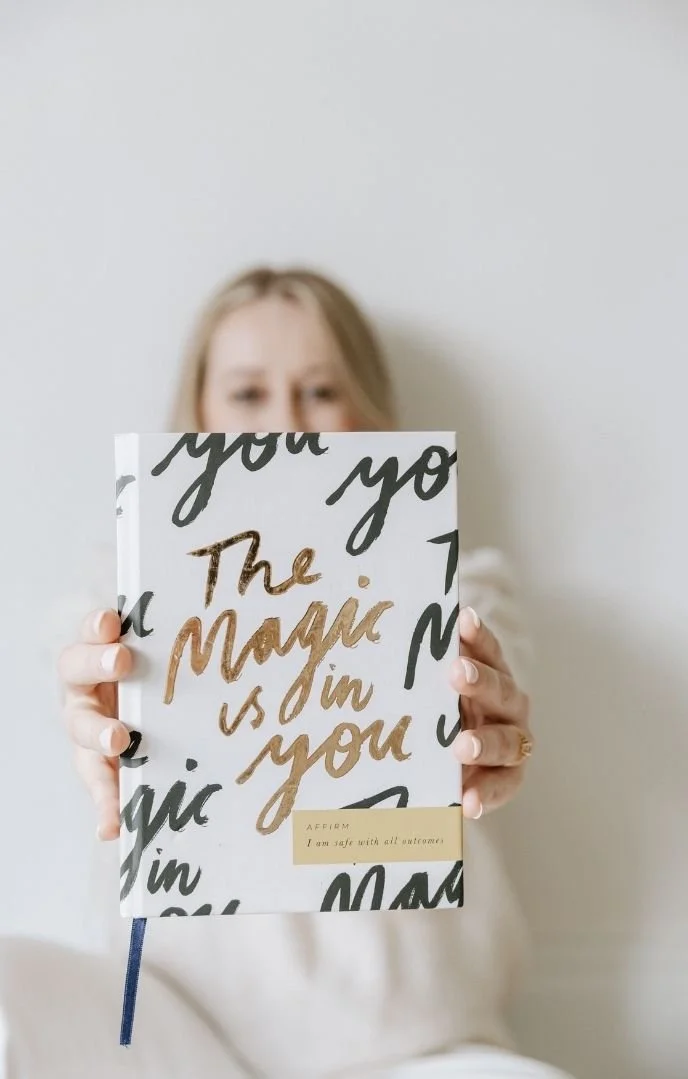 A woman holding a white book with gold and black lettering that says 'The magic is in you' and a small yellow label that says 'Affirm: I am safe with all outcomes,' standing against a light gray background.