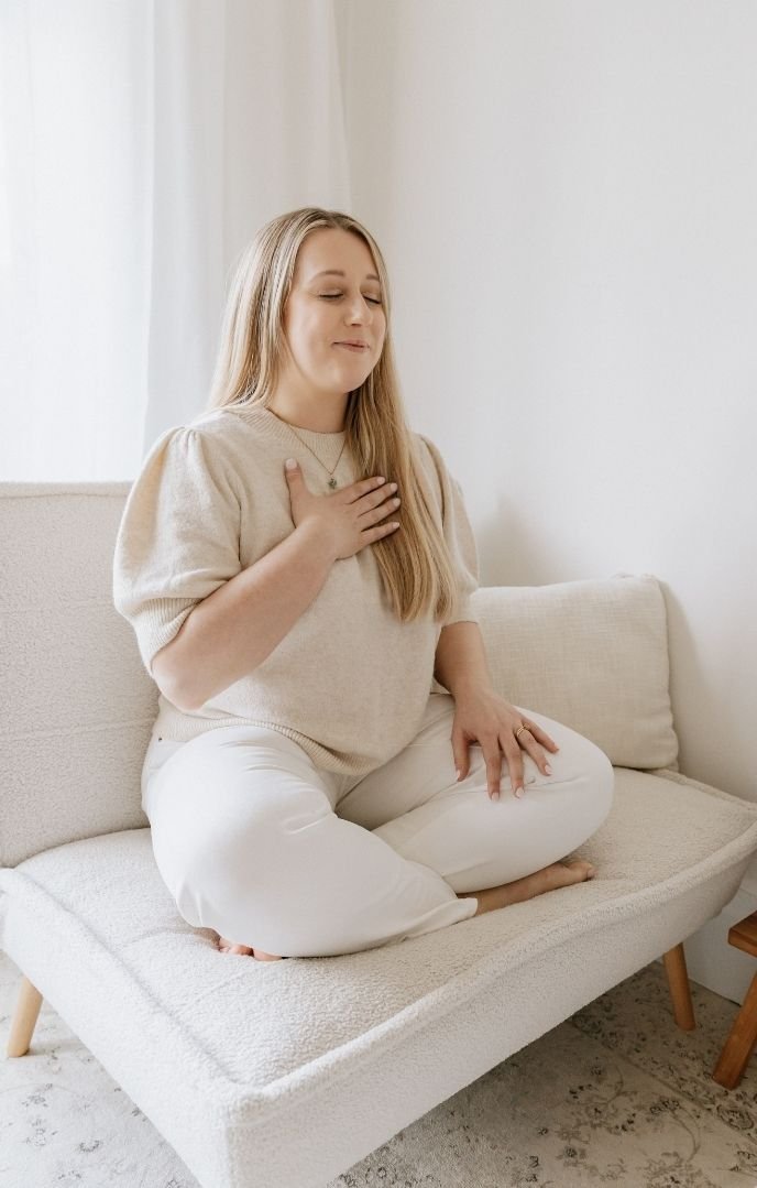 A young woman sitting cross-legged on a beige couch, placing her right hand on her chest, with a serene expression, in a bright room with white curtains.