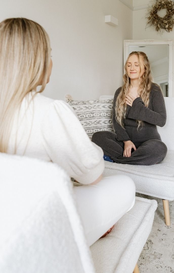 Two women are sitting in a cozy, well-lit room, engaged in a heartfelt conversation. One woman, with long blonde hair, is facing away from the camera wearing a white top. The other woman, with long wavy blonde hair, is sitting cross-legged on a sofa, wearing a dark gray outfit, and has her hand on her chest with a smile, indicating an emotional moment.