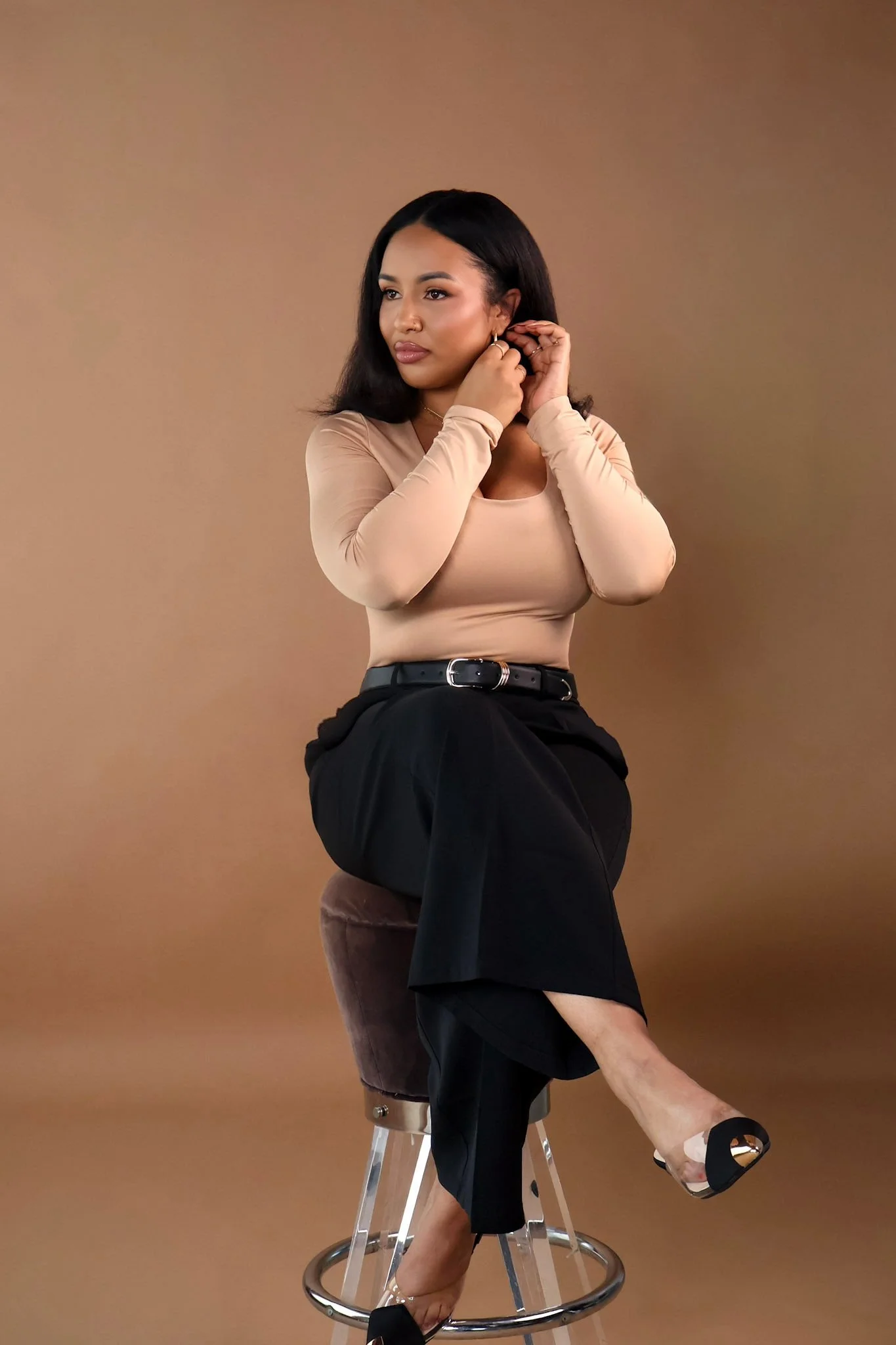 A woman with long dark hair in a beige top and black skirt sitting on a brown counter stool, adjusting her earring, with a tan background.