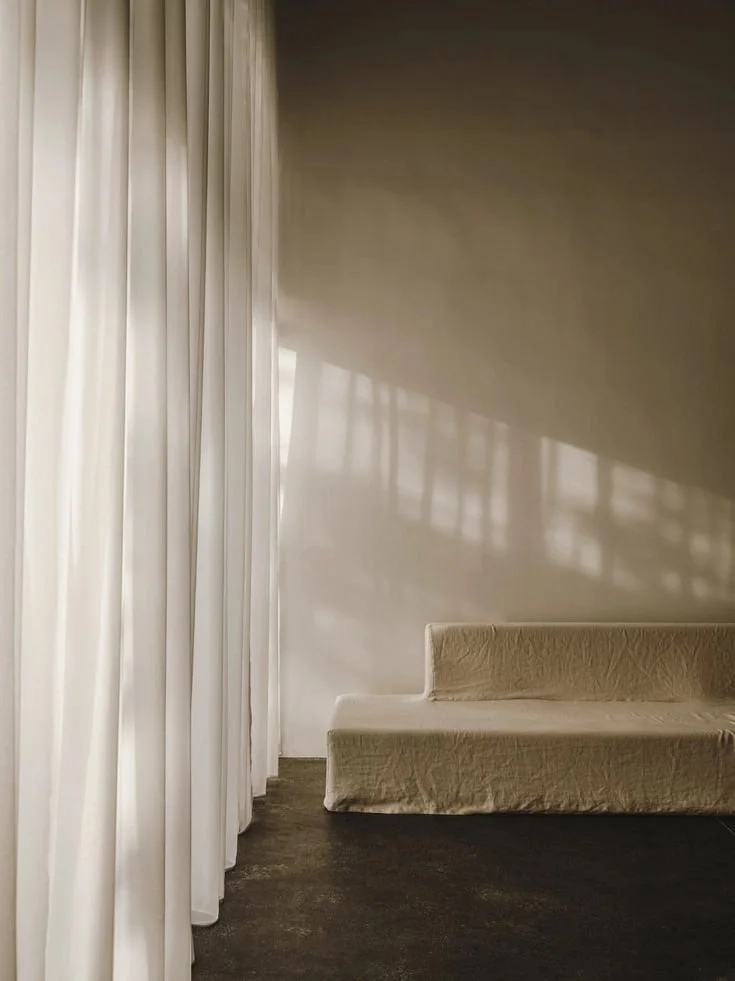 A minimalistic room with white curtains on the left and a cream-colored sofa on the right, with soft shadows cast on the white wall.