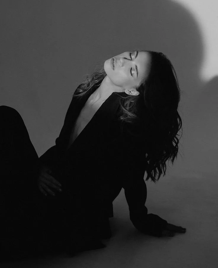 Black and white photo of a woman with long wavy hair, sitting with her eyes closed and head tilted back, wearing a dark blazer, against a plain background with a light spot.