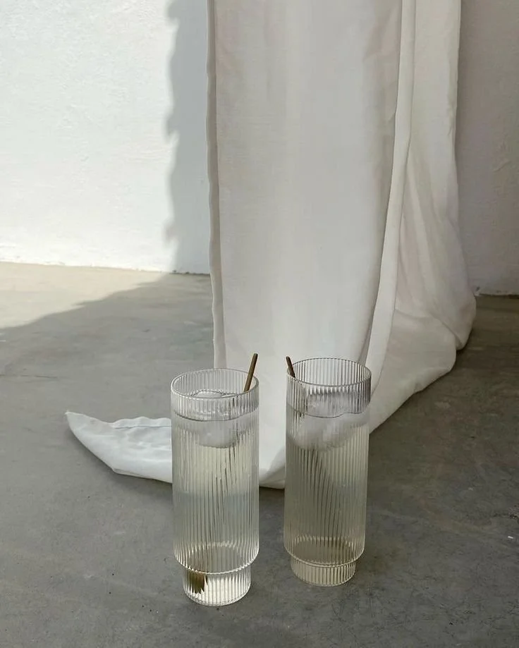 Two tall, ribbed glasses filled with water and ice, each with a straw, placed on a concrete floor in front of a white fabric backdrop.