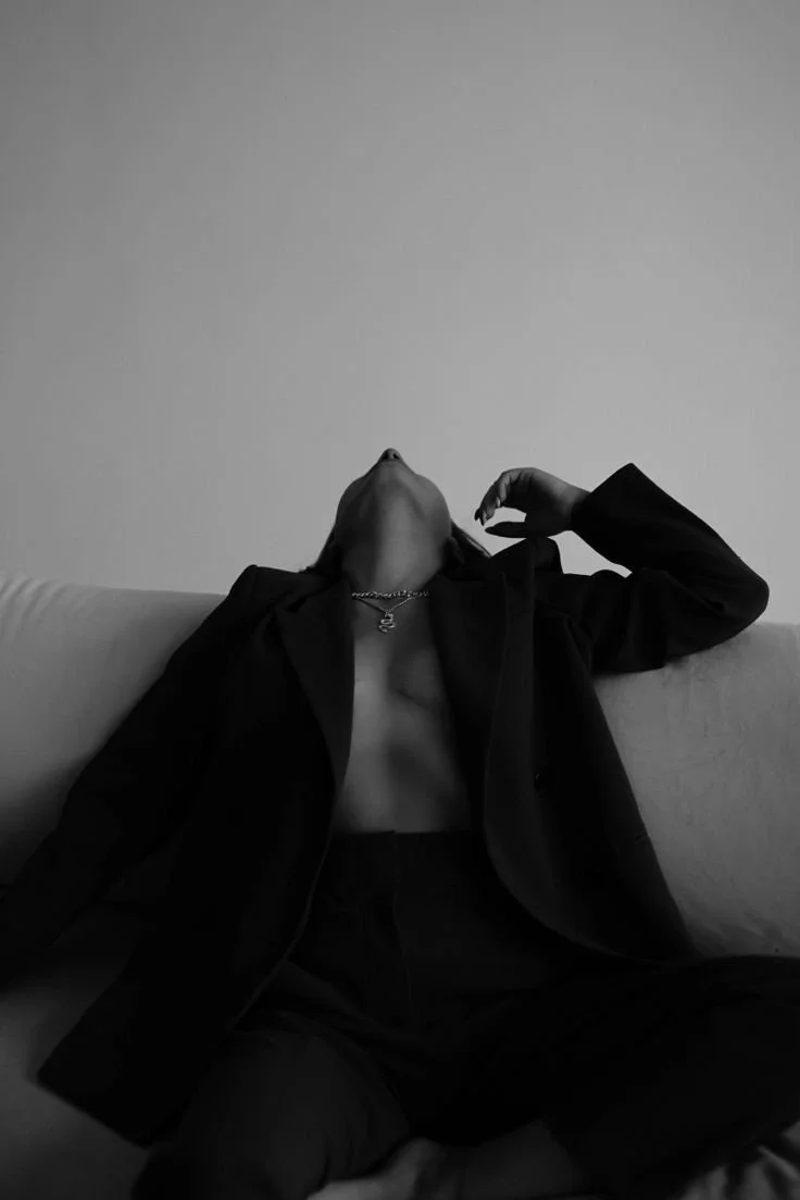 A person sitting on a couch, wearing a blazer and pants, with their head tilted back and eyes closed, in a black and white photo.