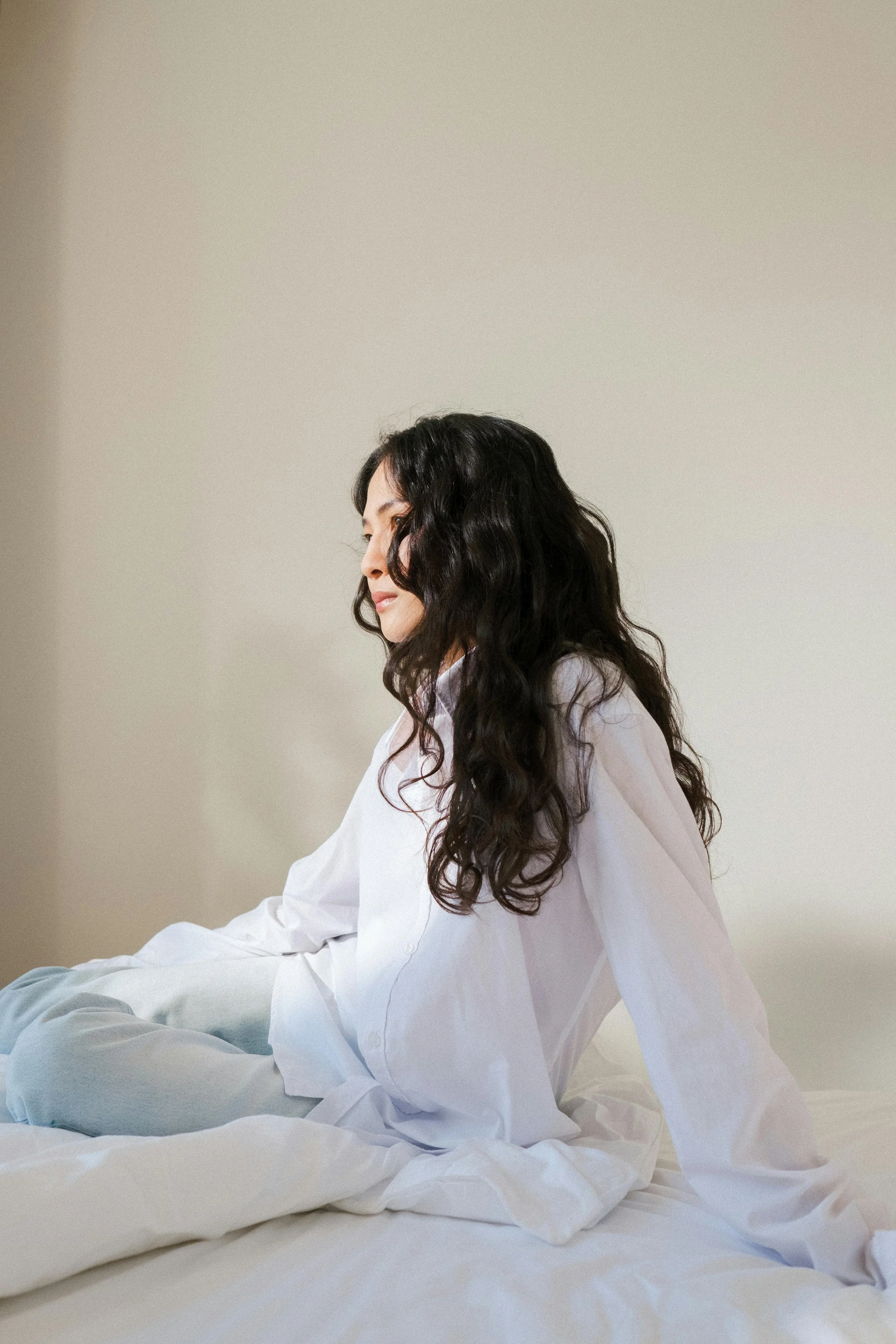 A woman with long, wavy black hair sitting on a bed, wearing a white shirt, with a neutral expression in a minimalist room with a plain beige wall.