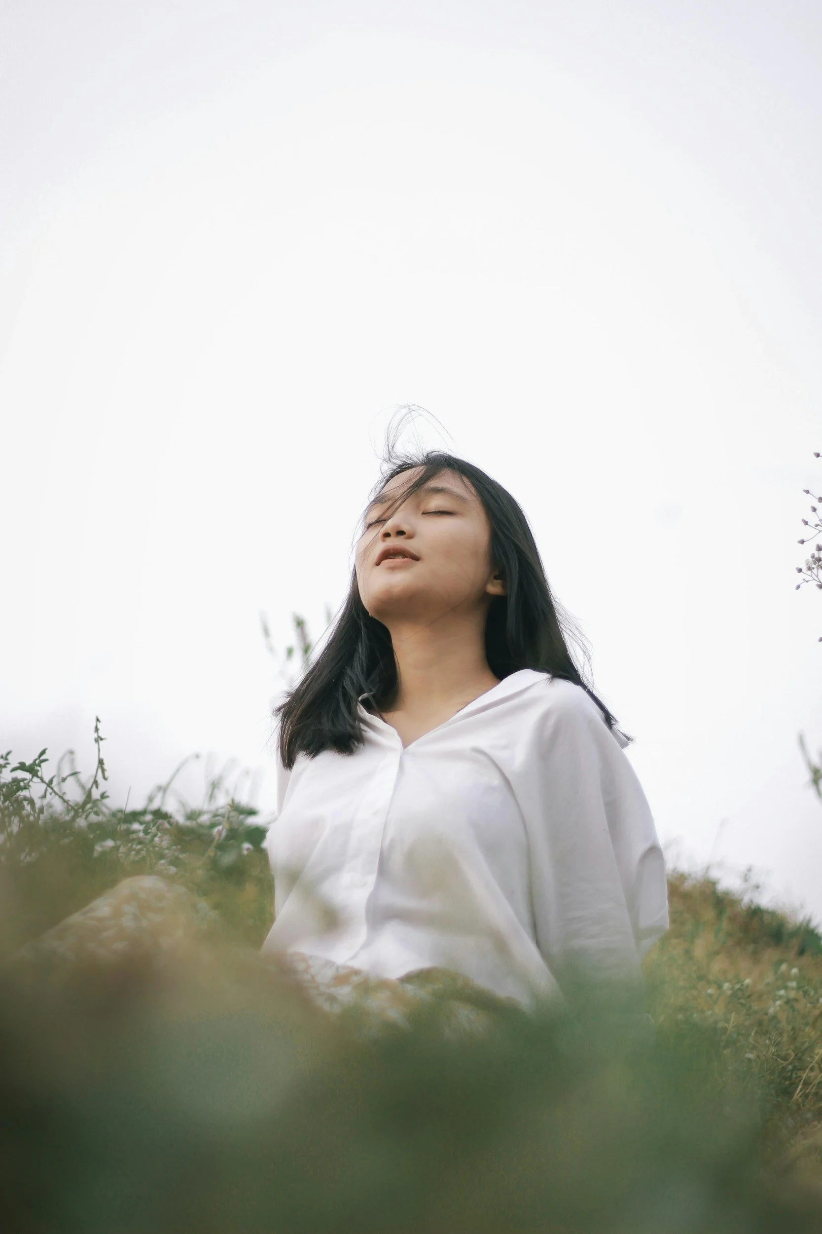 A woman with black hair in a white shirt standing outdoors with her eyes closed, surrounded by greenery, against an overcast sky.