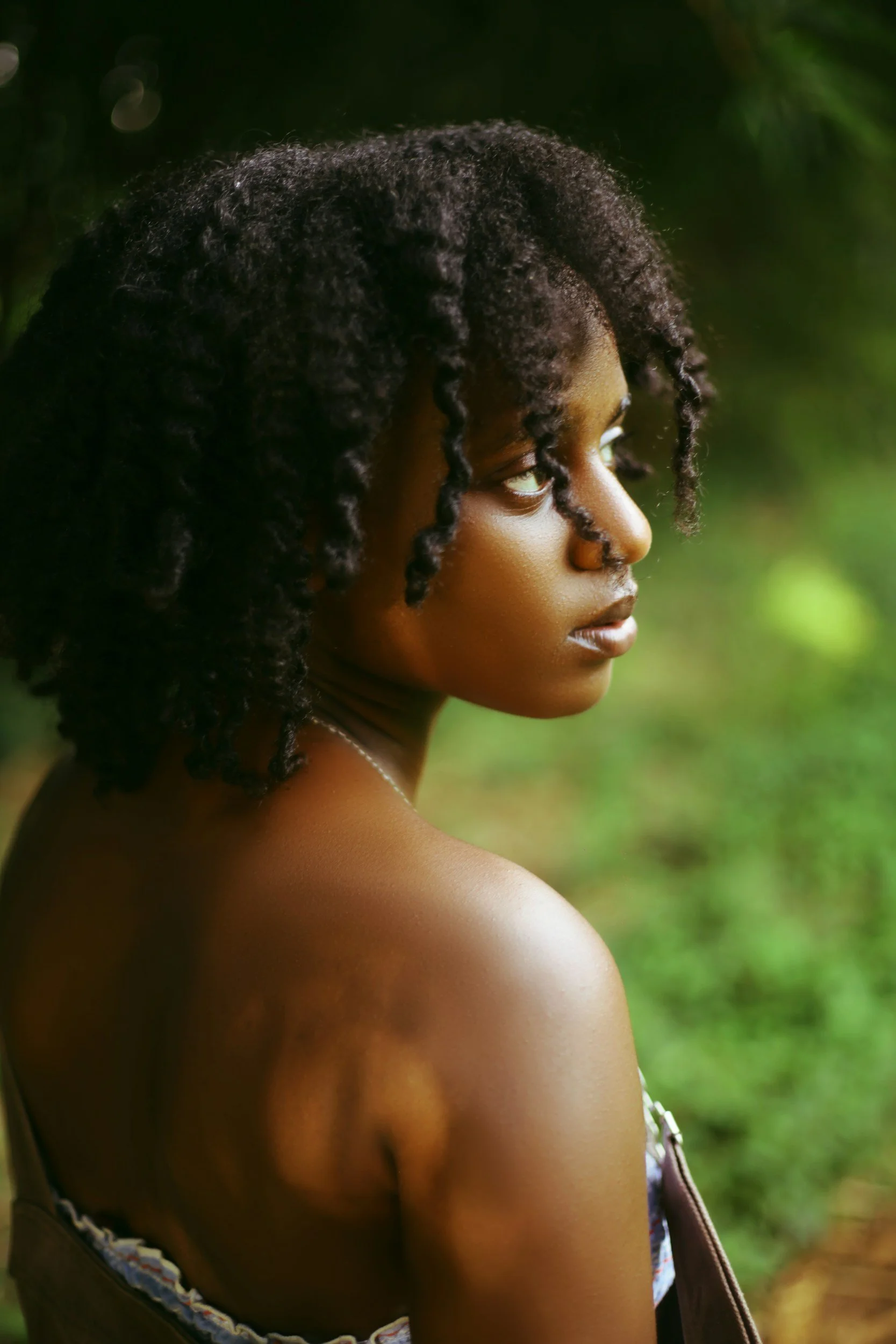 A young woman with dark, textured, natural hair looking over her shoulder outdoors surrounded by greenery.