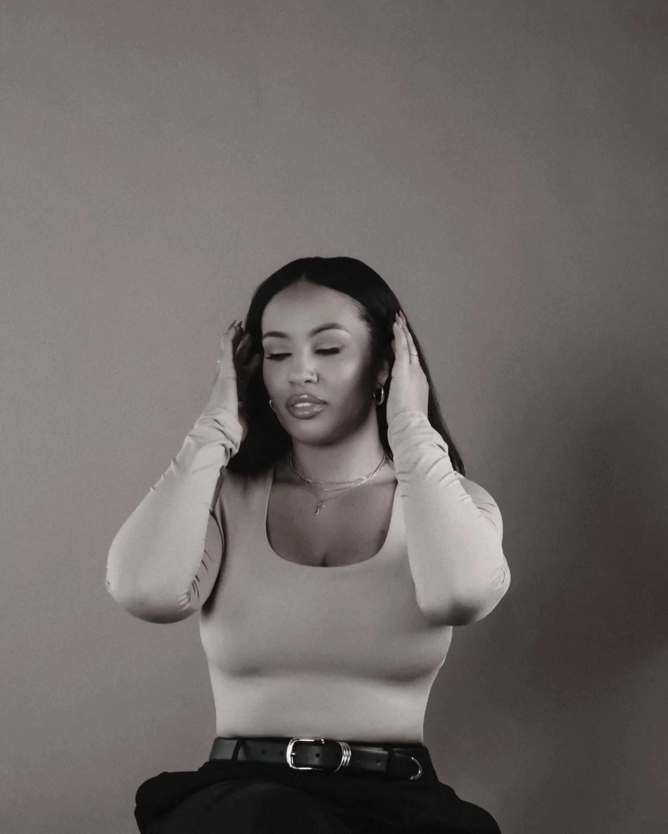 Woman with long dark hair holding her head with both hands, closed eyes, wearing a fitted long-sleeve top, necklaces, and hoop earrings, against a plain background.
