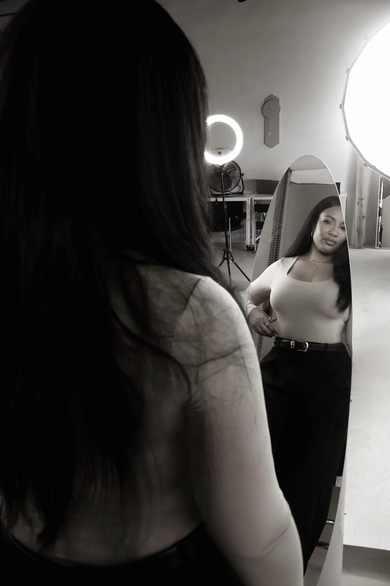 A woman looking into a standing mirror, with professional studio lighting and equipment in the background.