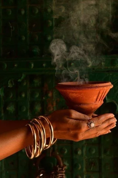 A person's hand holding a small, decorated clay pot with smoke rising from it. The hand is adorned with gold bangles and a ring, and the background features a green, ornate surface.