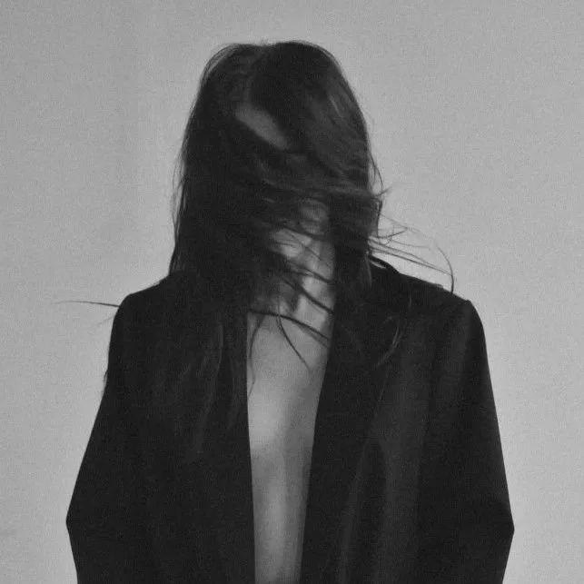 Black and white photo of a woman with long, messy hair covering her face, wearing an open jacket with no shirt underneath.