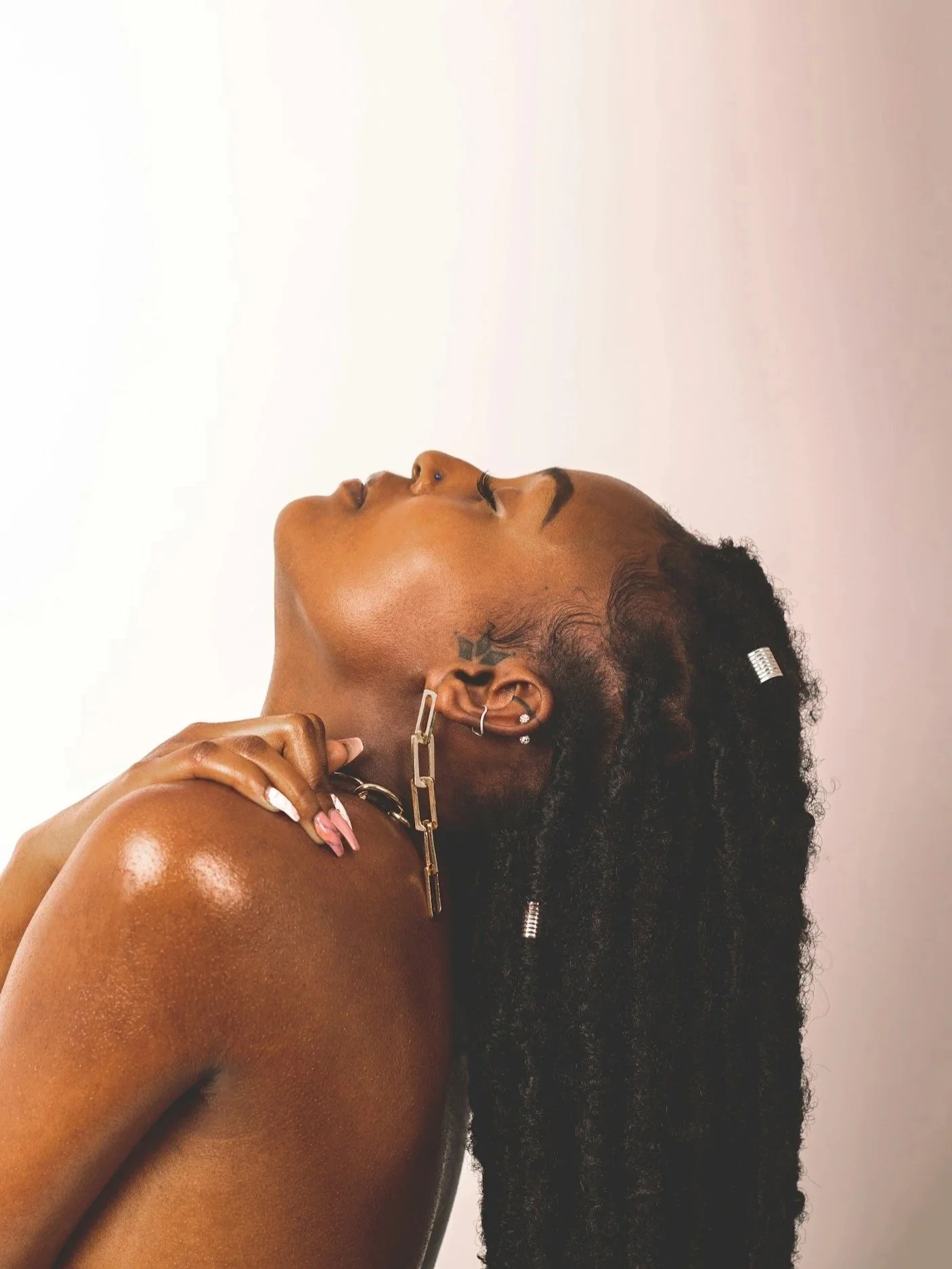 A woman with dark skin and textured black hair styled with hair clips, wearing large metal jewelry, leans her head back with eyes closed against a plain background.
