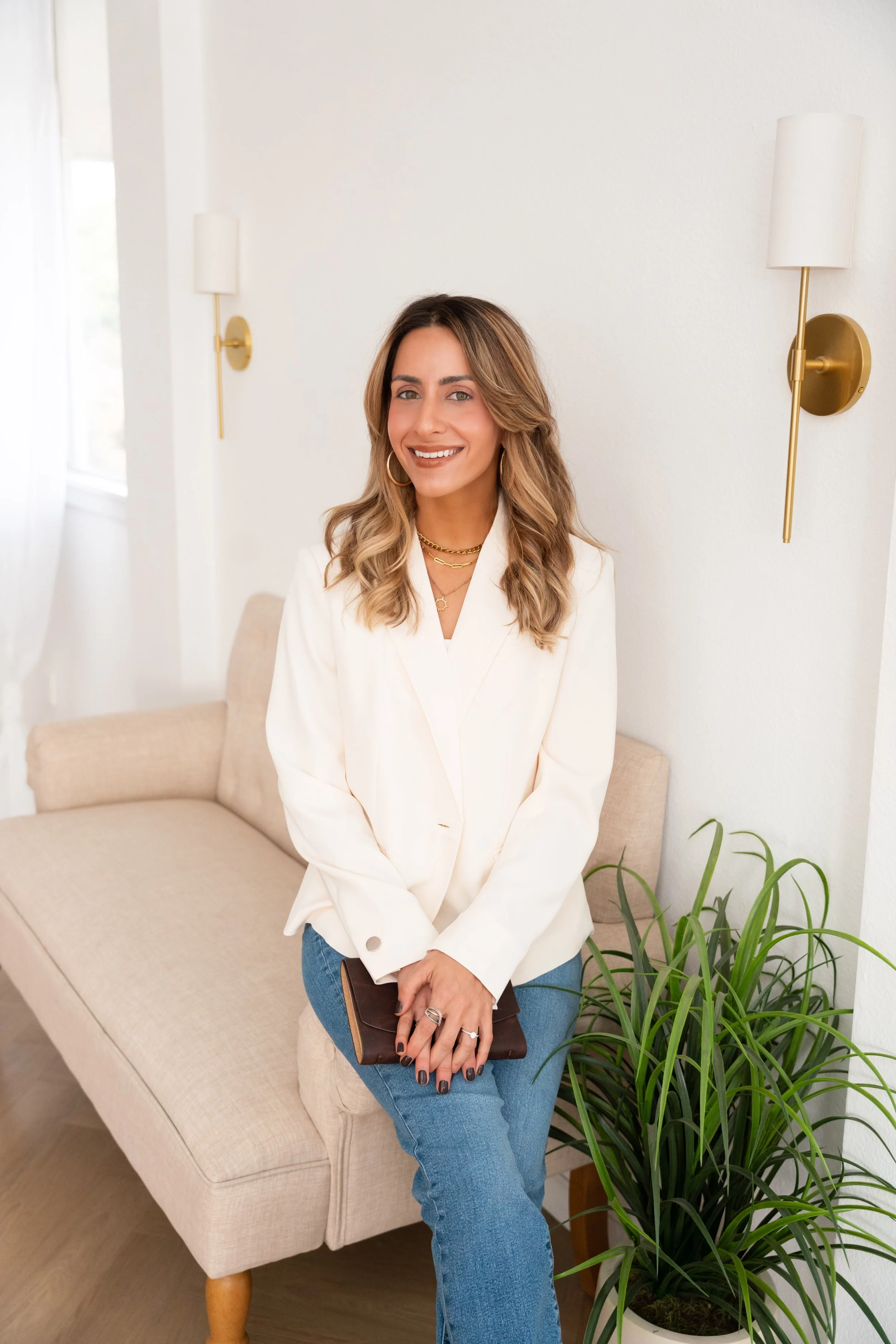 A woman sitting on a beige sofa, smiling, with gold wall sconces and a green plant nearby, in a bright, modern room.