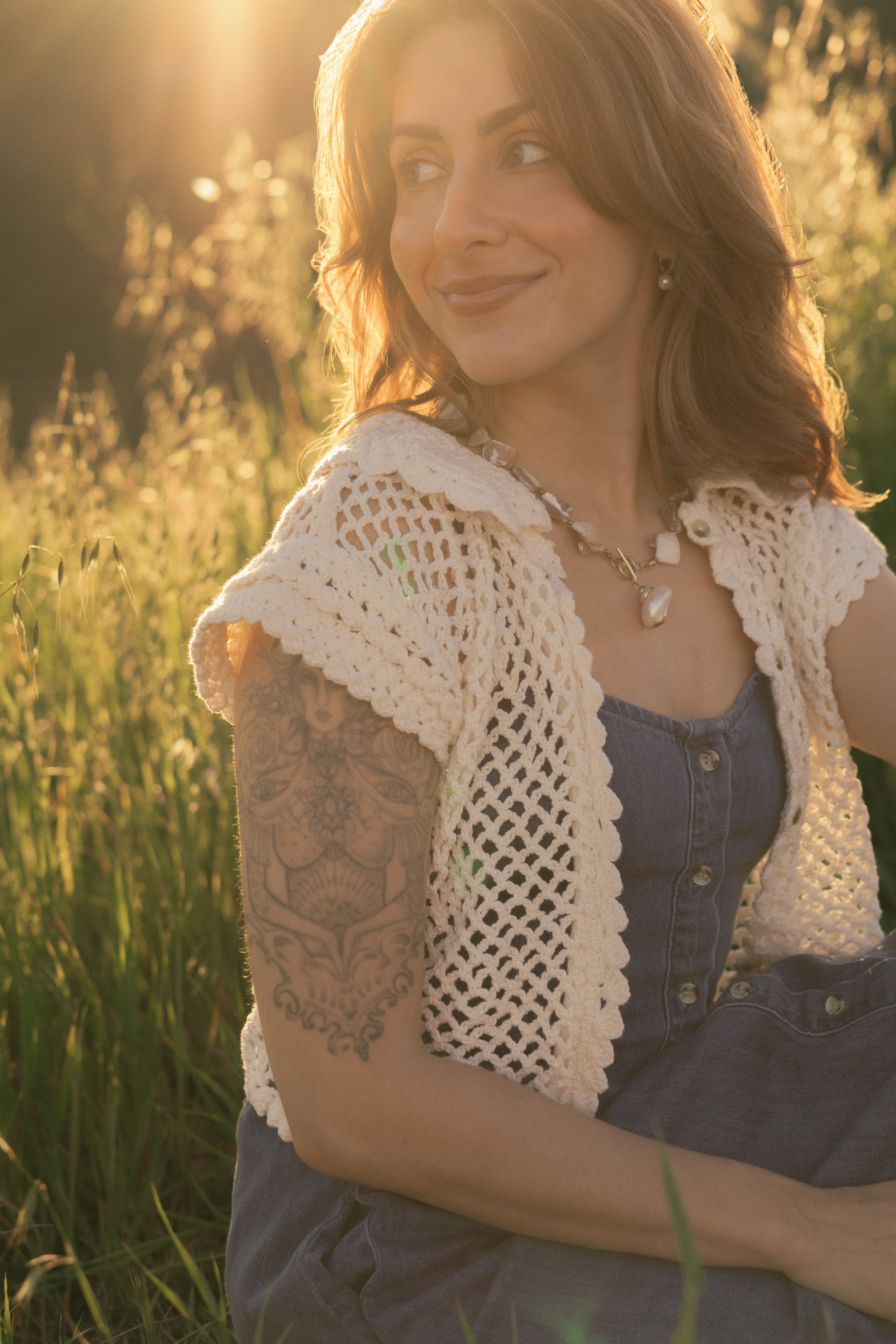 A woman sitting in a field during sunset, smiling while looking to the side. She has shoulder-length hair, a tattoo on her arm, and is wearing a crocheted top, a necklace, and earrings.