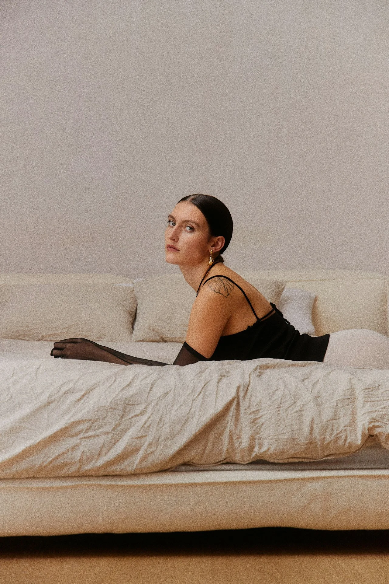 A woman with dark hair and tattoos, wearing a black dress and sheer gloves, sitting on a bed with a neutral expression, in a minimalistic room.