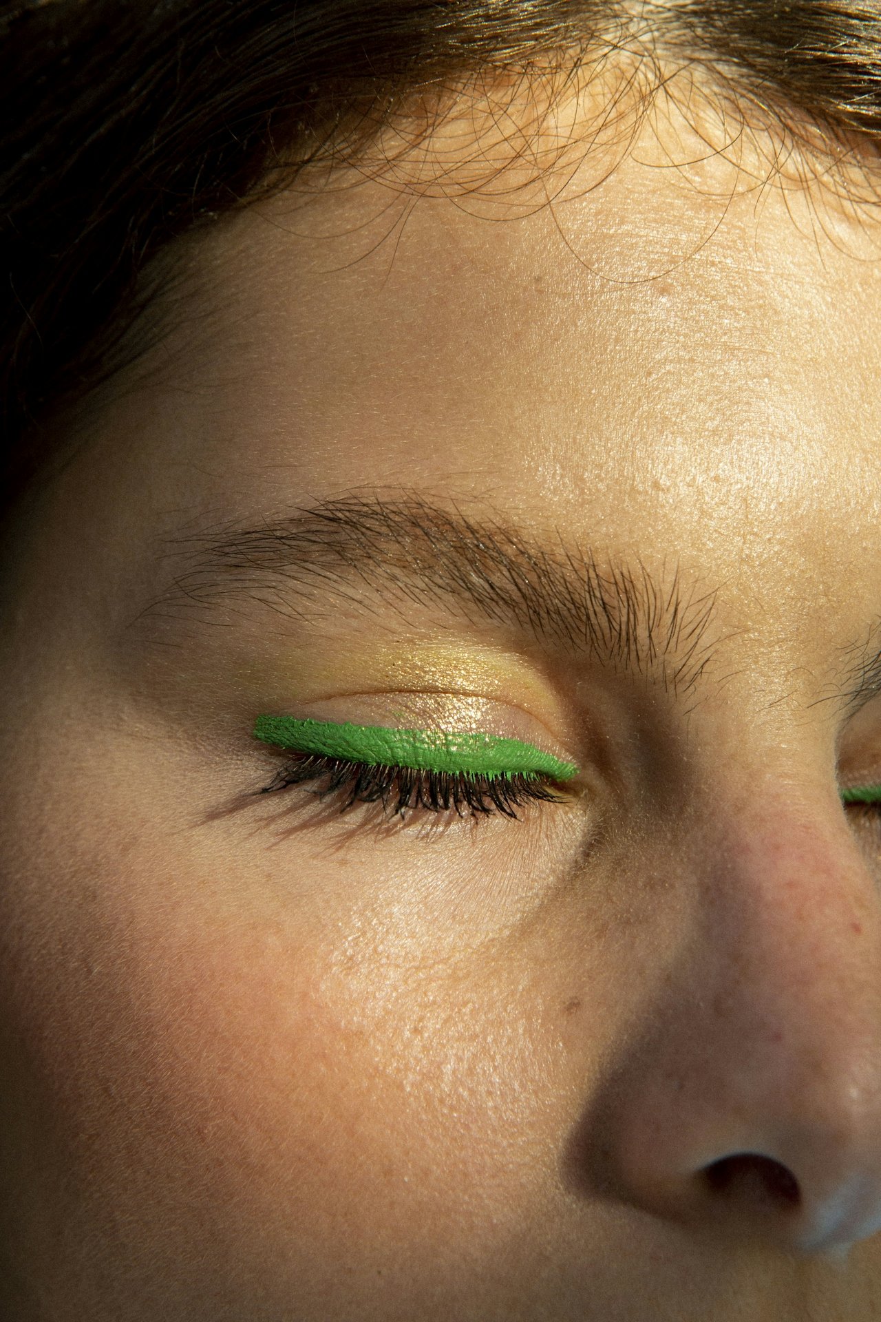Close-up of a person's face with closed eyes, wearing bright green eyeliner.