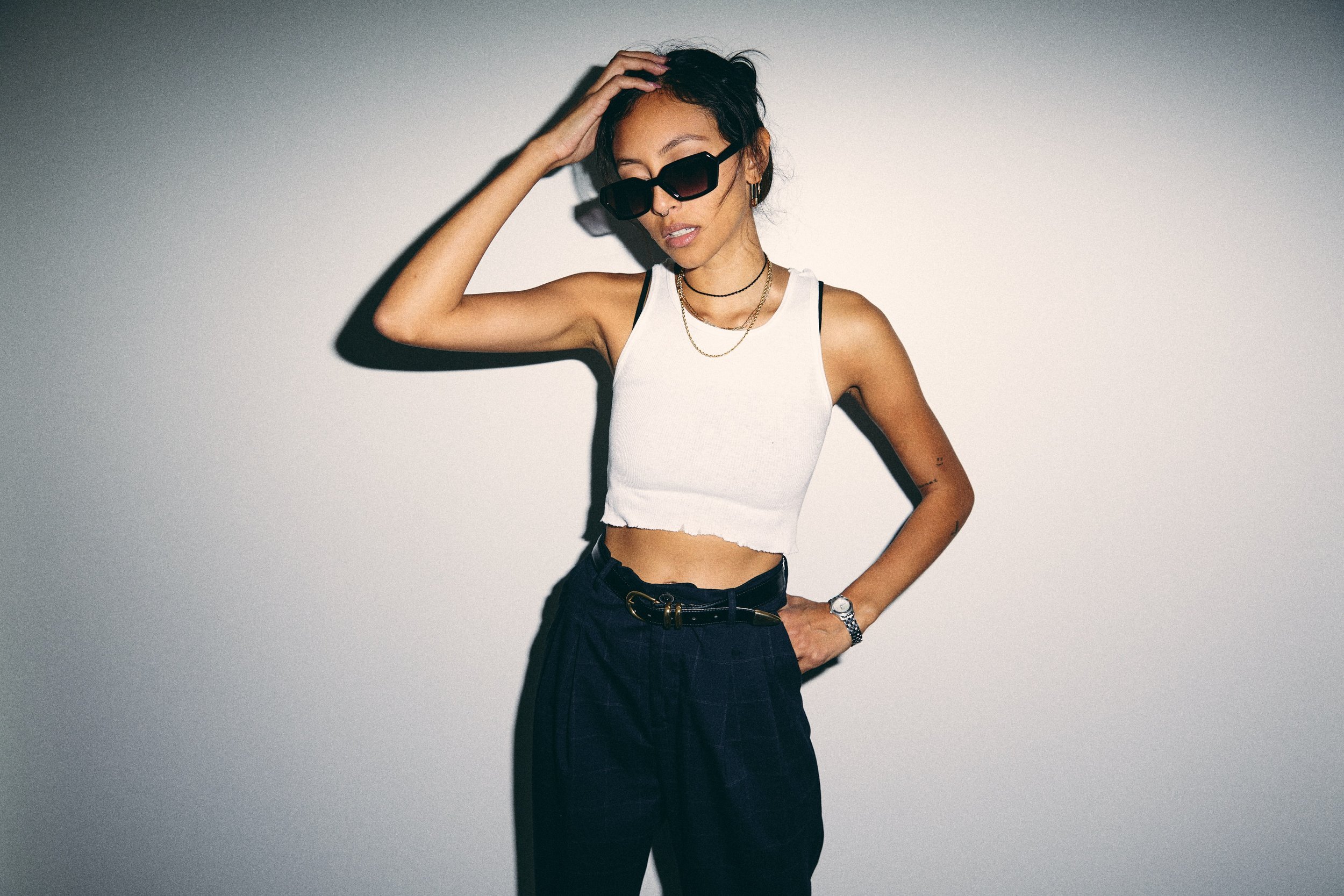 A young woman with dark hair wearing sunglasses, a white cropped tank top, black high-waisted pants, and accessories, standing against a plain light background.