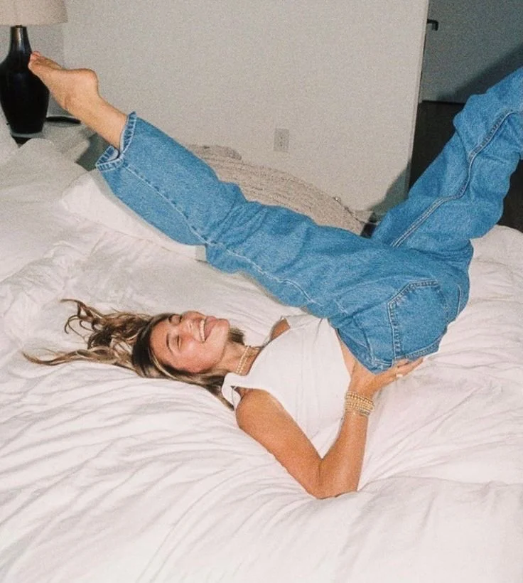 A woman lying on a bed with her legs raised and resting on the headboard, smiling with eyes closed, wearing a white sleeveless top, blue jeans, and a bracelet.
