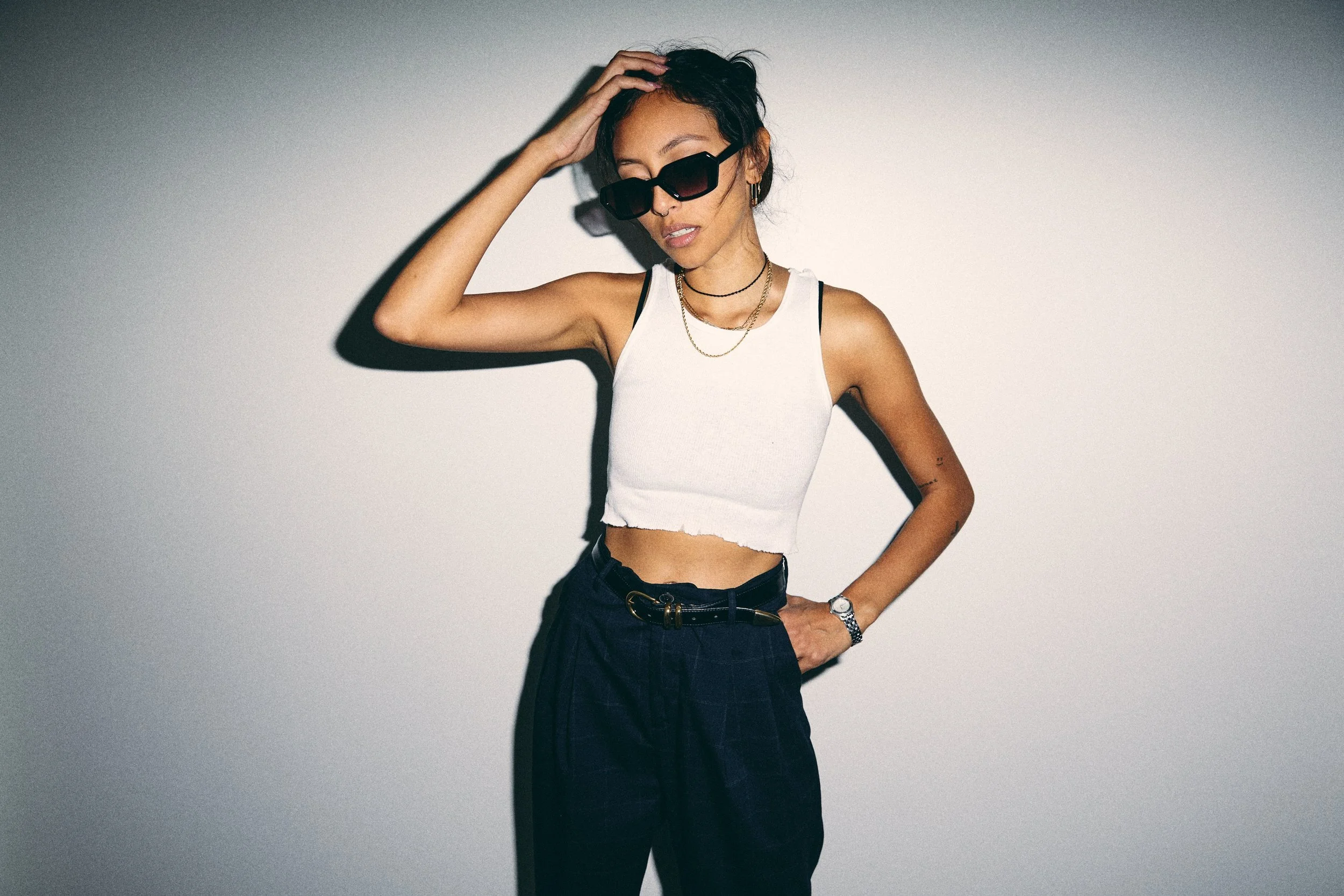 A woman wearing black sunglasses, a white crop top, dark high-waisted pants with a belt, layered necklaces, a watch, and earrings, posing against a plain wall with her hand on her head.