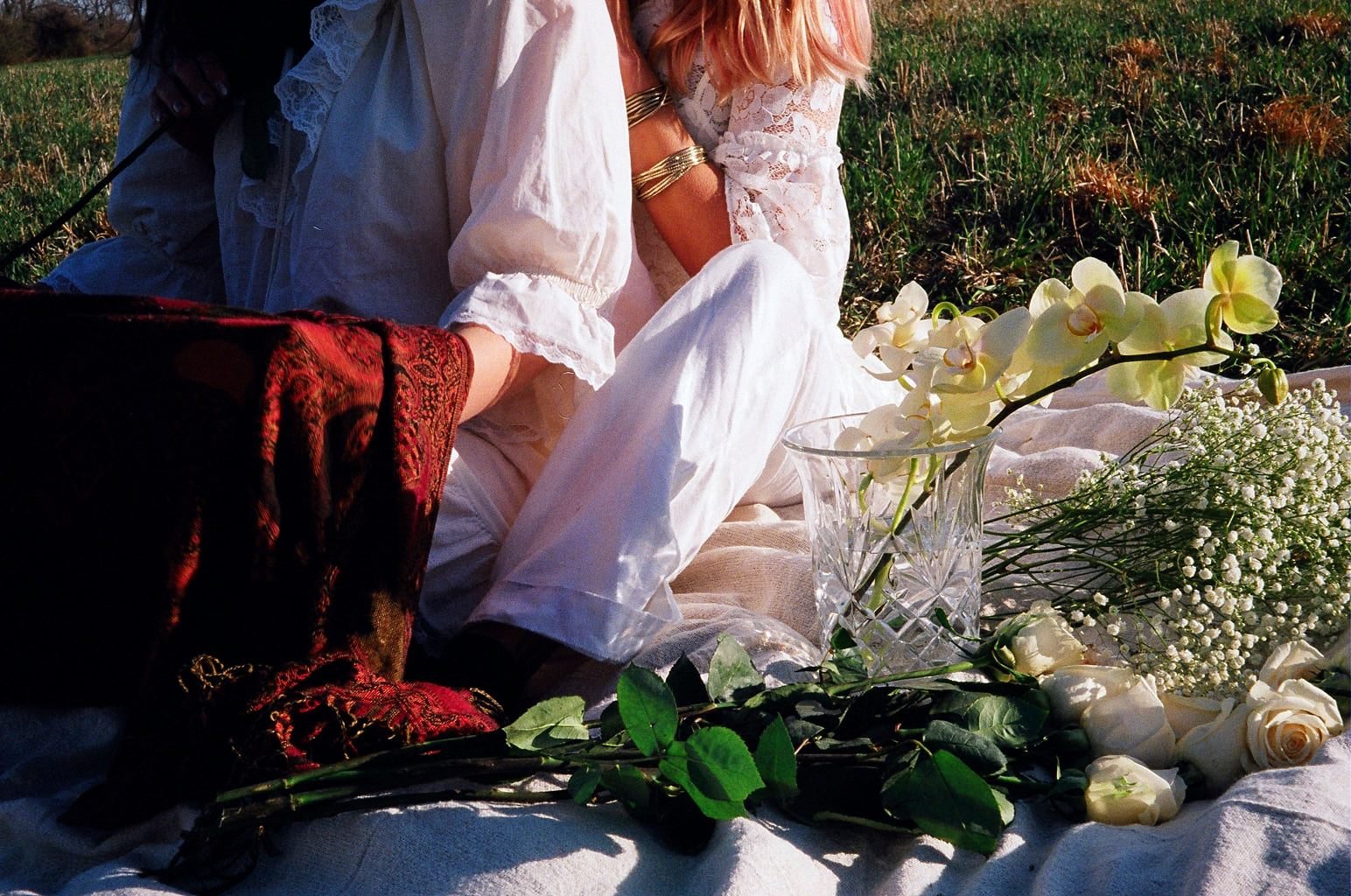 A person dressed in white sitting outdoors on grass with flowers and a vase around them.