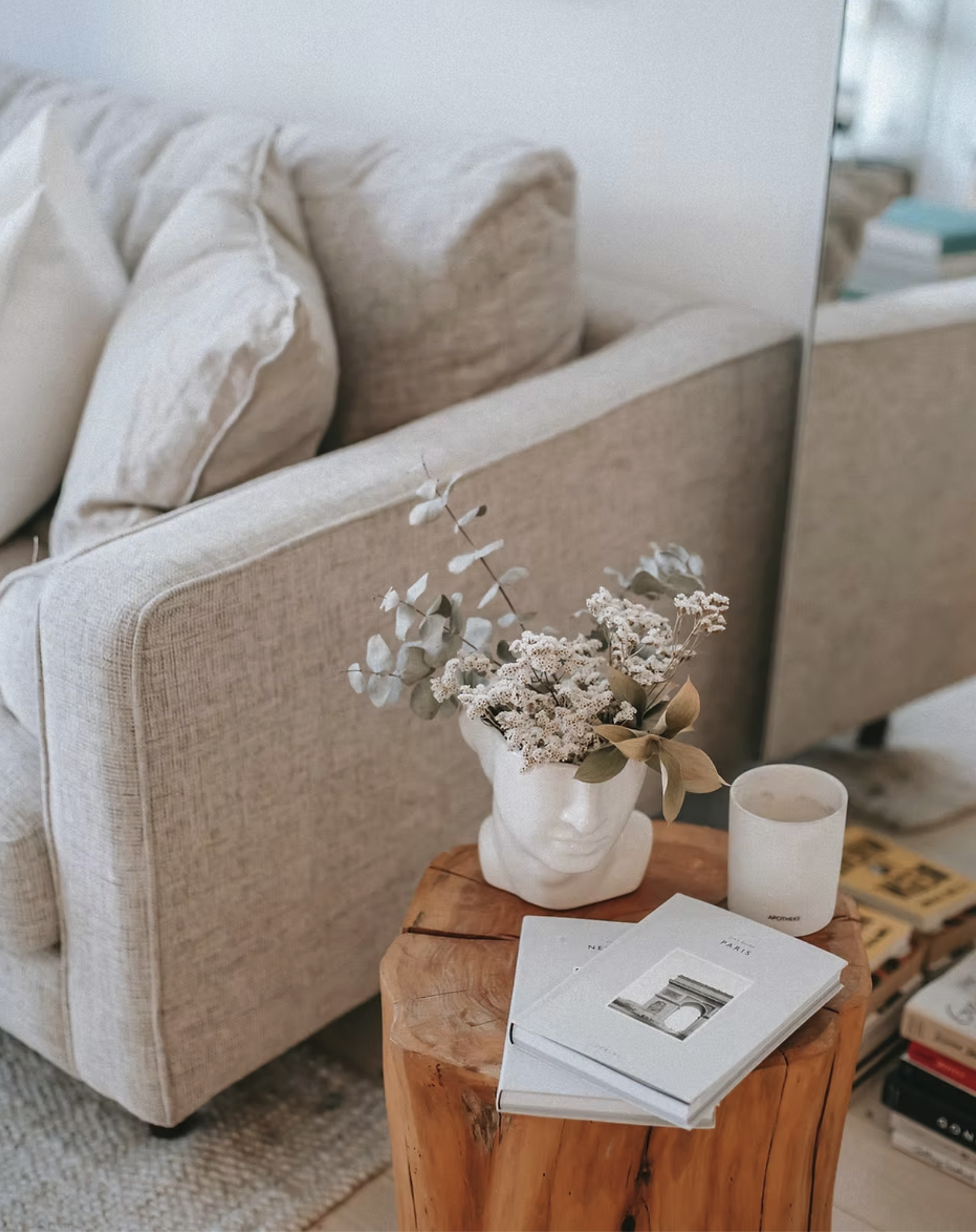 A cozy living room corner with a beige sofa, decorative pillows, a wooden side table with a white vase holding dried flowers, a white candle, and magazines.