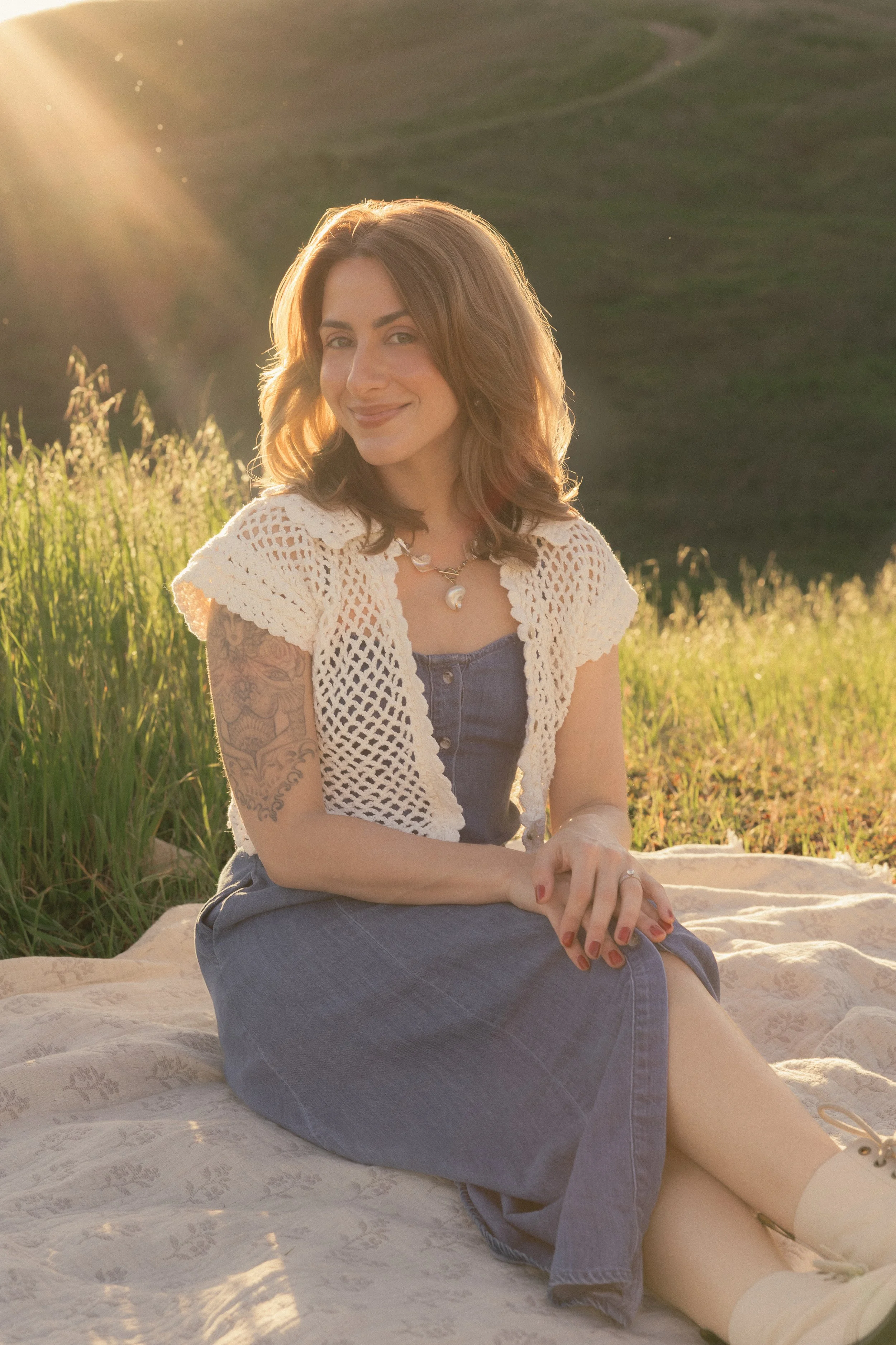 A woman with shoulder-length brown hair sitting outdoors on a blanket during sunset, wearing a denim dress, a white crochet cardigan, and a necklace, with a tattooed arm and a gentle smile.