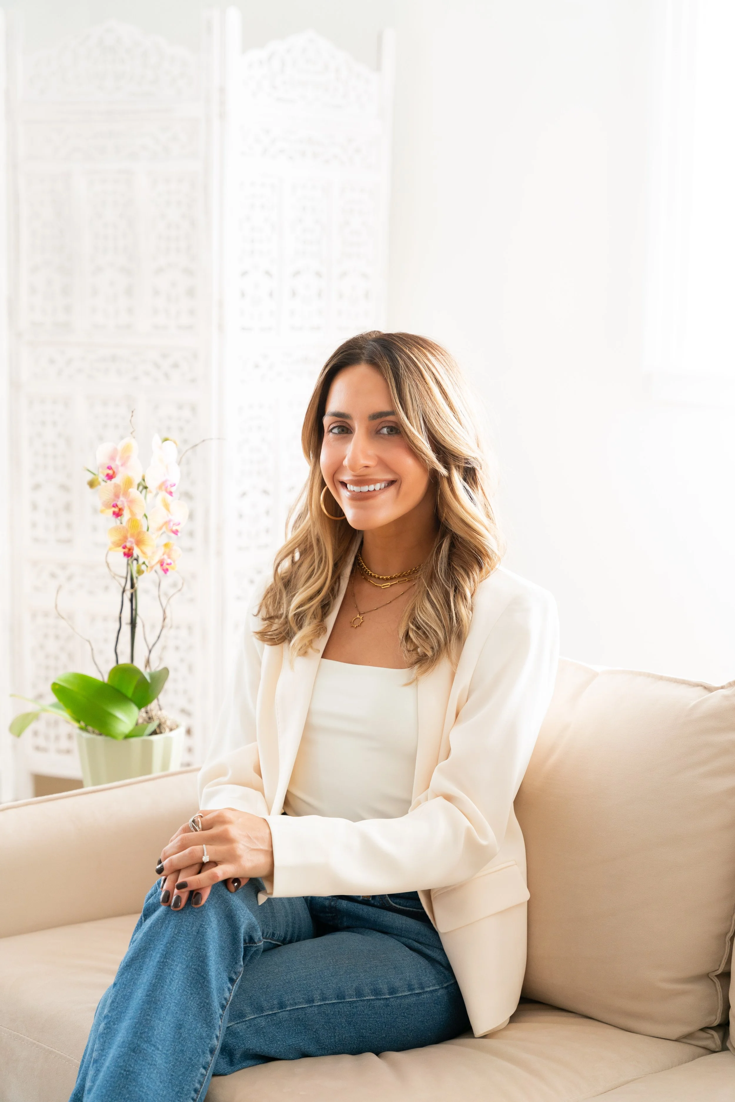 A woman with shoulder-length wavy brown hair, wearing a cream-colored blazer over a white top, sitting on a beige couch. She has gold jewelry, including hoop earrings, layered necklaces, and rings. The background features a white decorative screen and an orchid plant in a pot. Bright natural light illuminates the room.