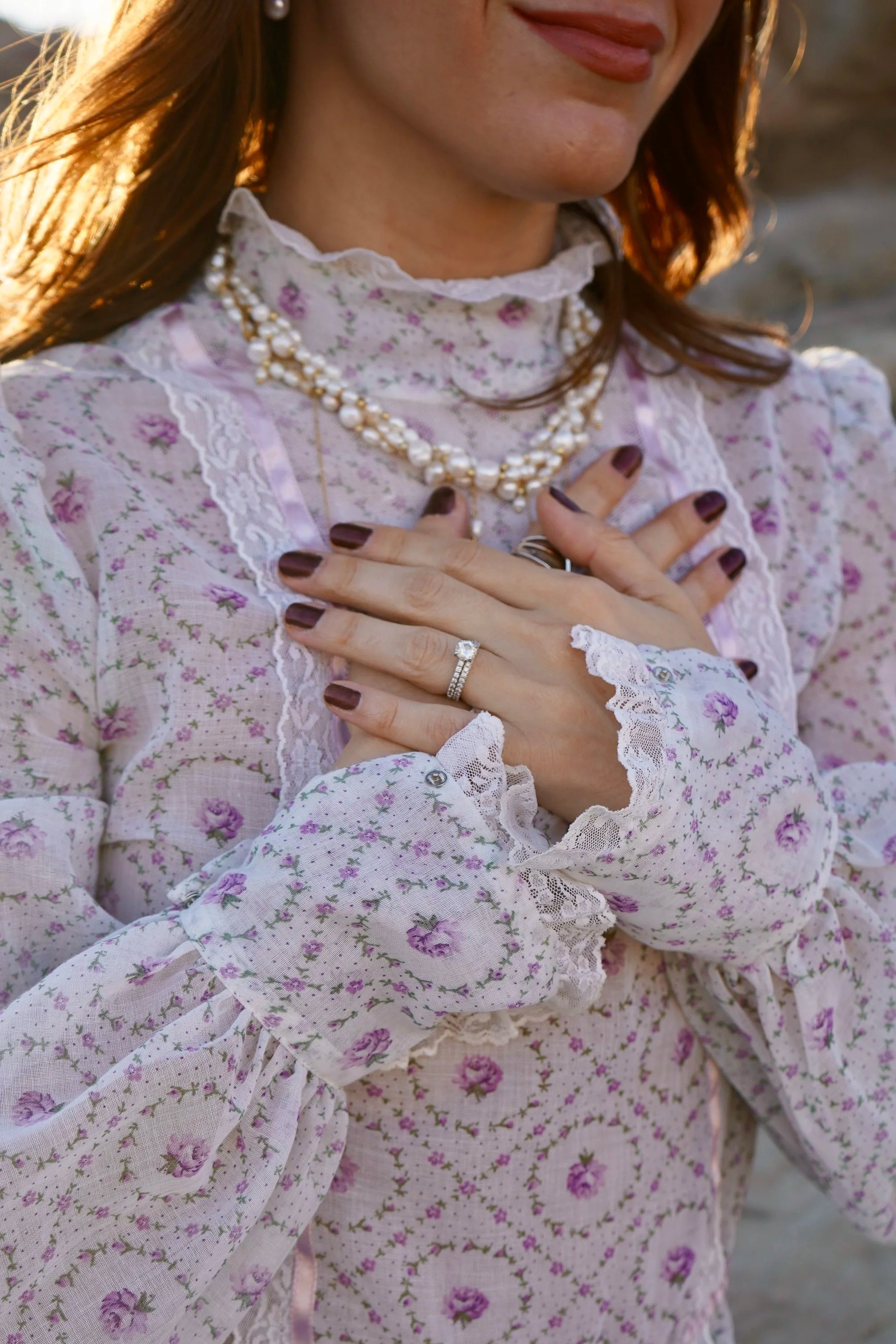 Close-up of a woman wearing a floral vintage dress, double-strand pearl necklace, and ring, with her hands crossed over her chest, palms down.