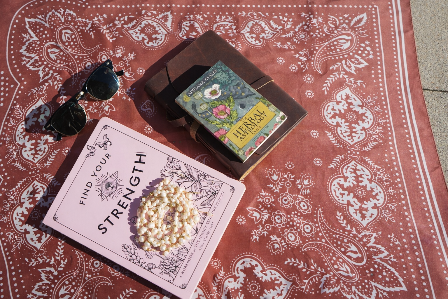 A red bandana with white floral and paisley patterns, placed on a surface with various items on top. These items include black sunglasses, a pink book titled 'Find Your Strength', a necklace with white pearls, a closed brown journal, and a small book titled 'The Herbal Astrology' by Adriana Ayales.