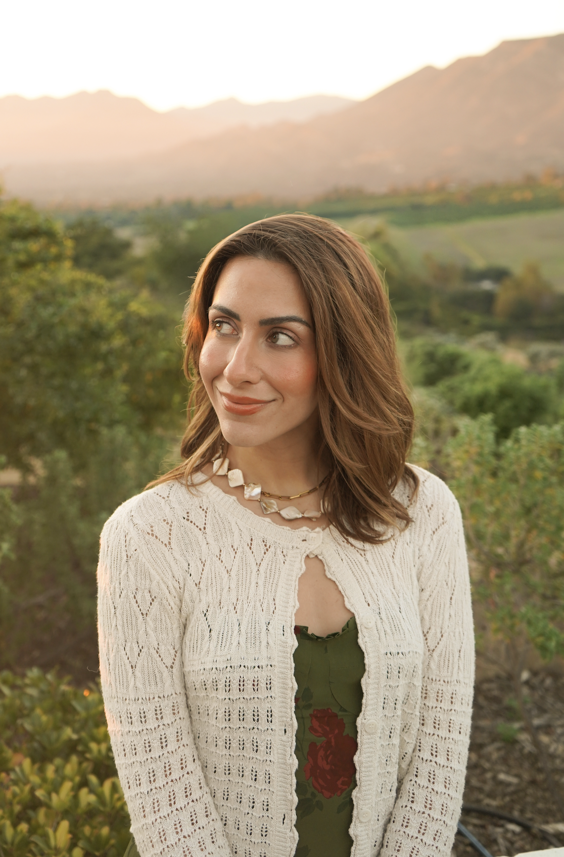A woman with shoulder-length brown hair wearing a white knit cardigan and green dress with red floral pattern, standing outdoors at sunset with a mountainous landscape and lush greenery in the background.