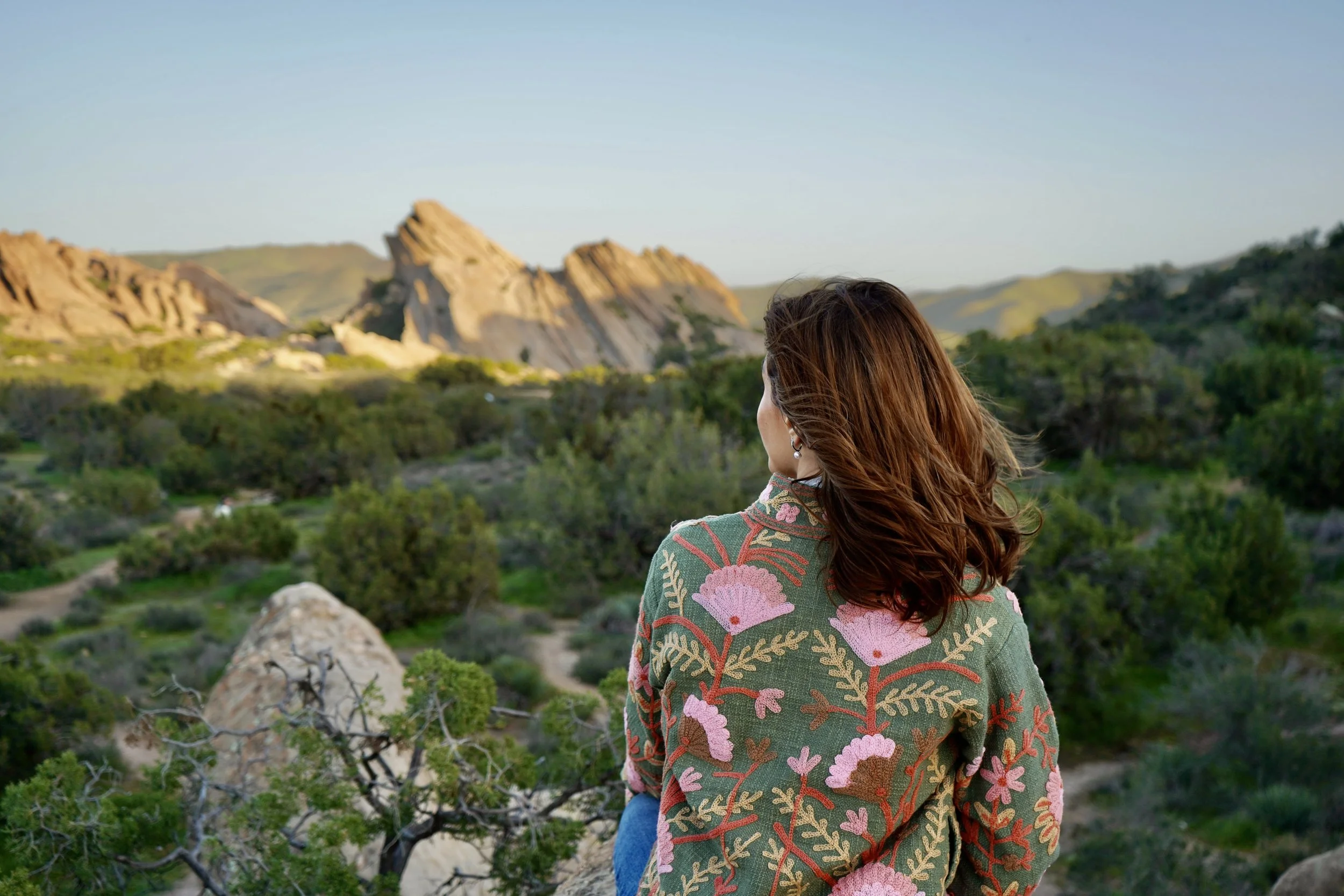 A woman with brown hair wearing a green floral jacket looking at rocky formations and mountains in a scenic landscape during sunset.
