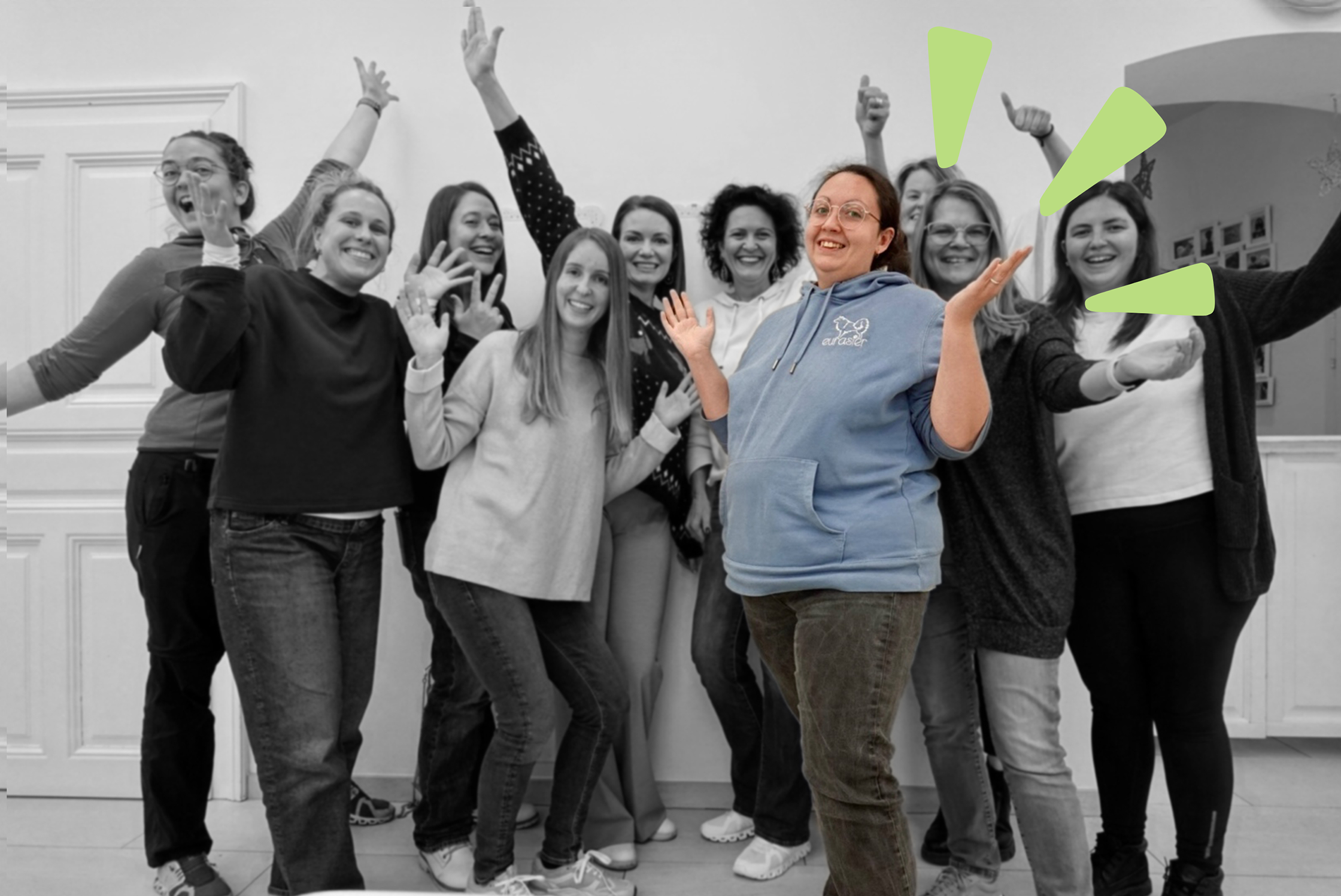 Eine Gruppe von Frauen, eine davon in Farbe und fröhlich, während die anderen im Hintergrund schwarz-weiß sind, die Frau erscheint in Farbe und lächelt mit erhobenen Armen.