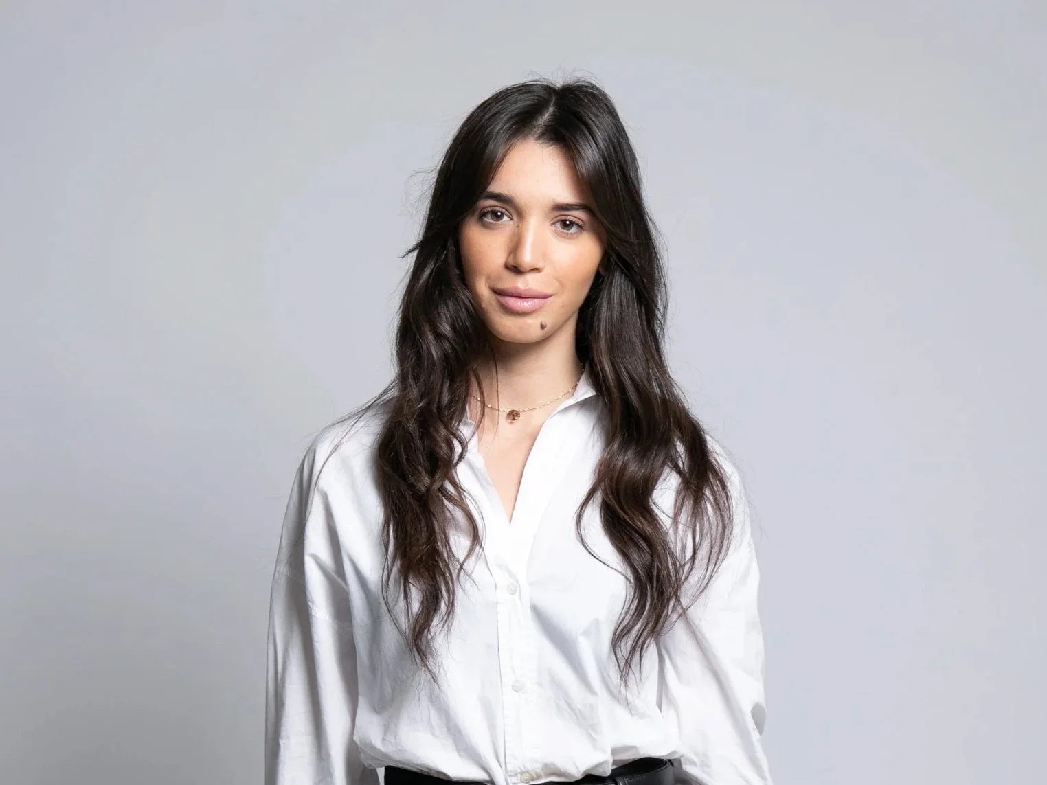 Une jeune femme aux cheveux longs, bruns, coiffés en vagues, portant une chemise blanche, se tenant contre un fond gris clair.