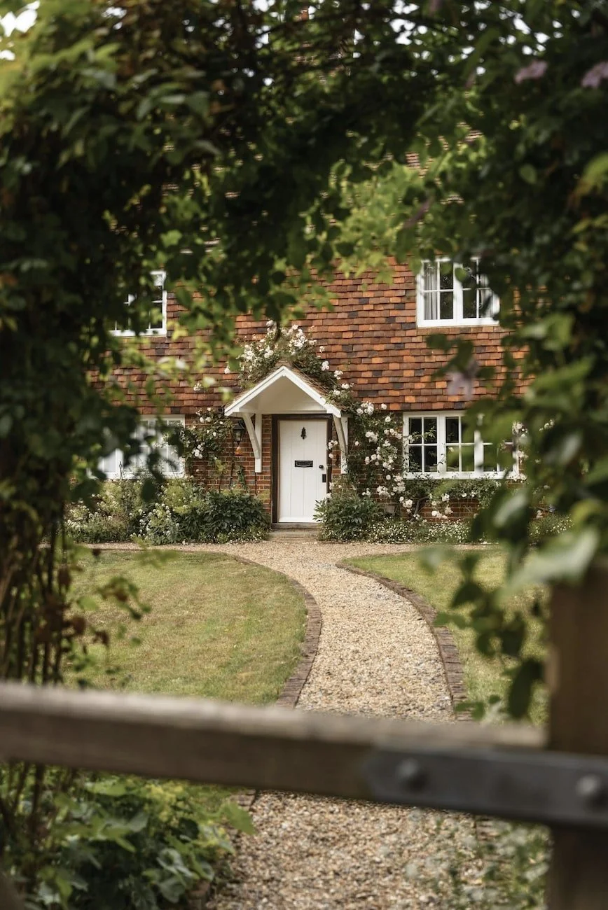 A brick house with white-framed windows, a white front door, and a small gabled porch. The house is partly obscured by green foliage in the foreground, with a curved gravel pathway leading to the door, and a well-maintained lawn.