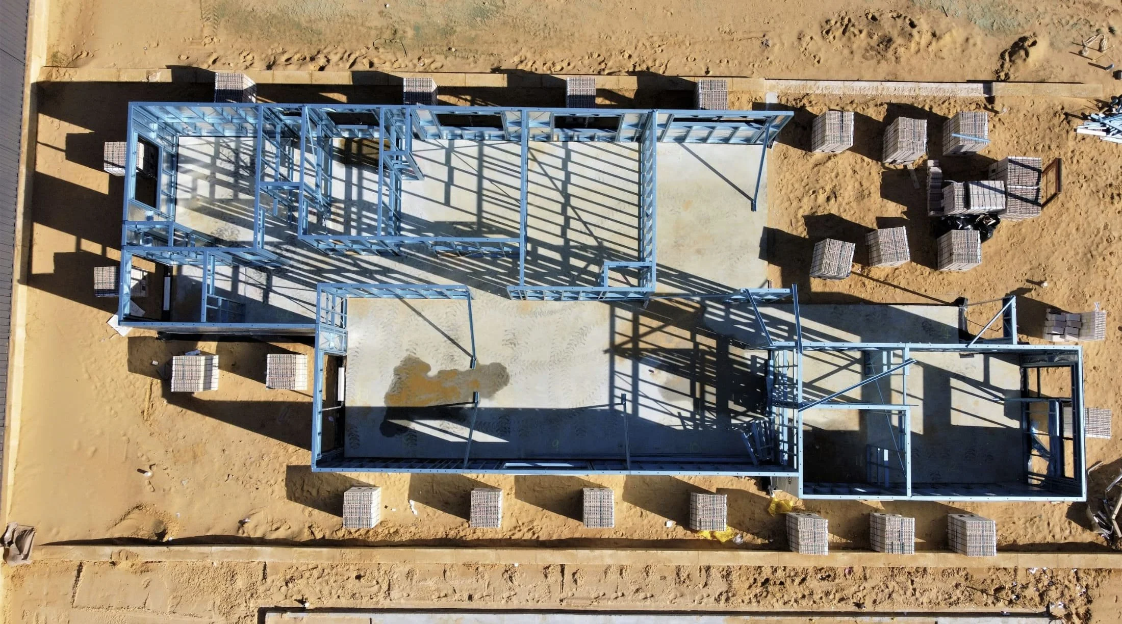 Aerial view of a construction site with steel framing for a building, surrounded by plastic-wrapped pallets of construction materials on sandy ground.
