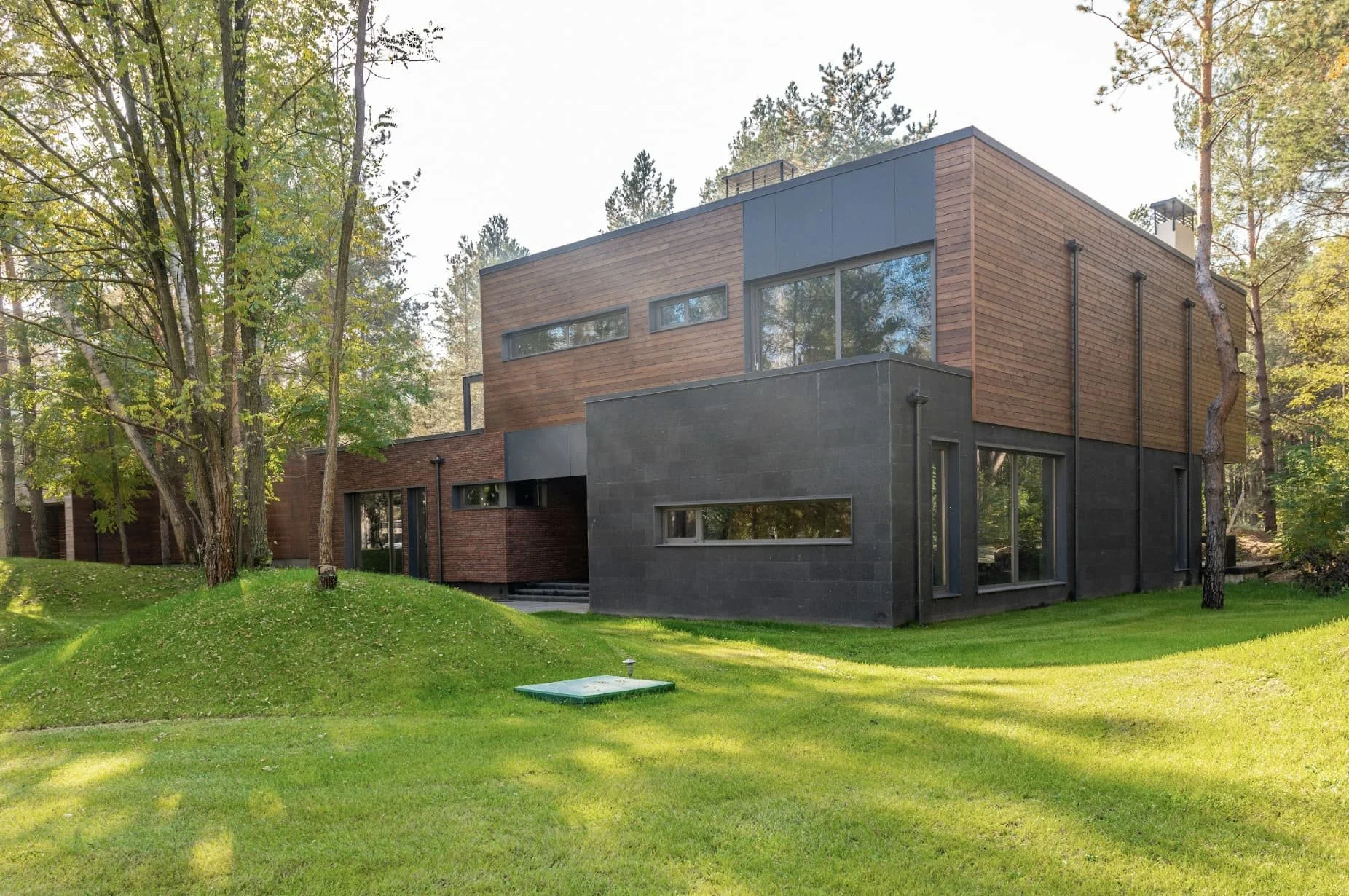 Modern multi-story house with black, brown, and wood exterior, surrounded by green lawn and tall trees.