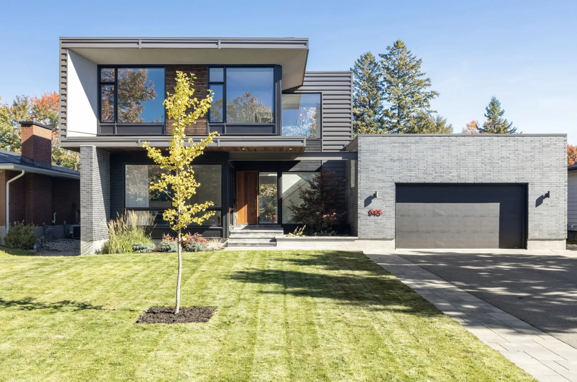 A modern two-story house with large glass windows, a gray brick facade, a garage, and a front yard with a small tree and well-maintained lawn.